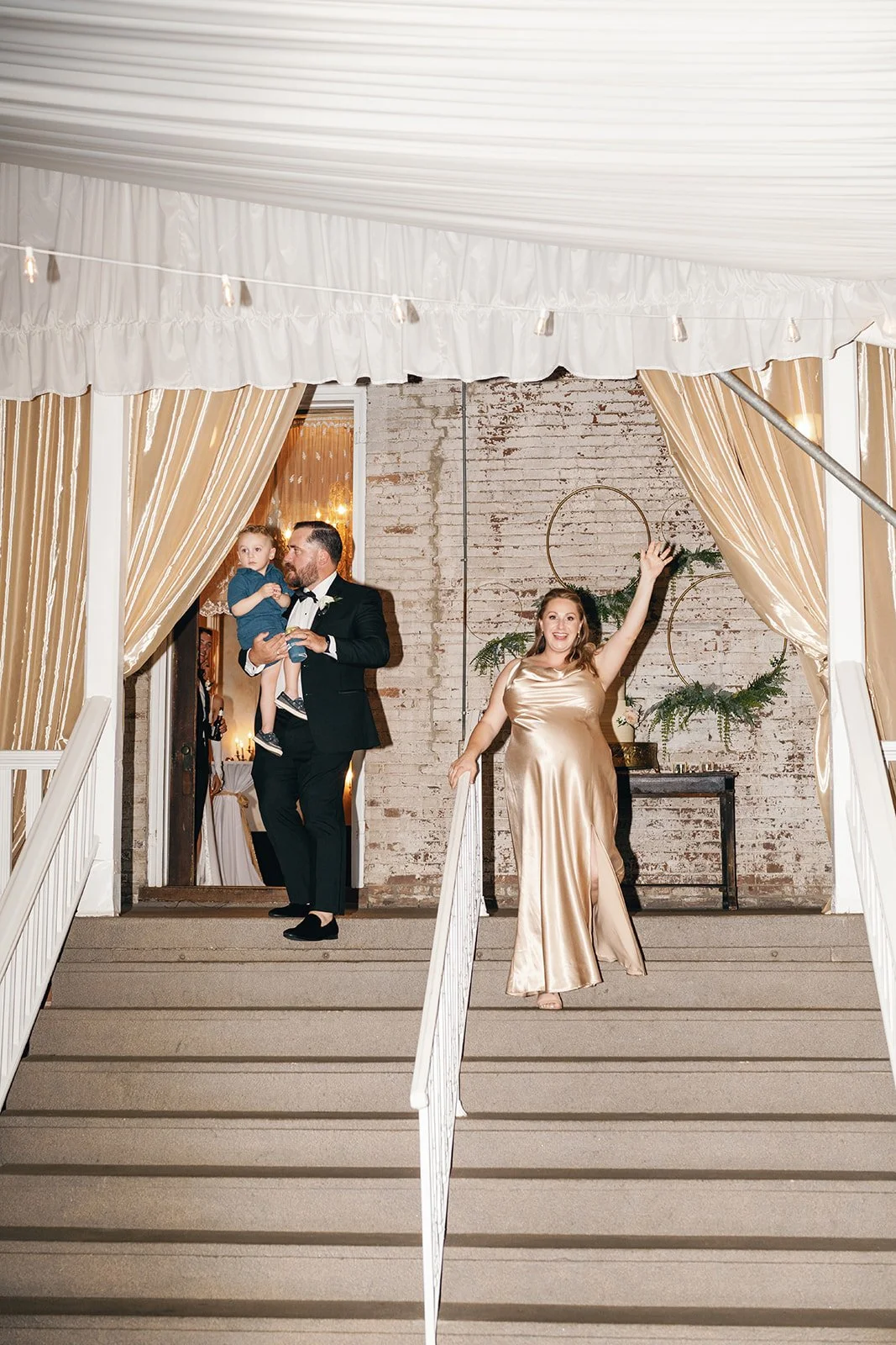 People standing on a staircase at a decorated venue, with a woman in a champagne-colored evening gown waving, a man in a tuxedo holding a young girl in a blue dress at the top of the stairs, and a brick wall with greenery and circular wall decor behi