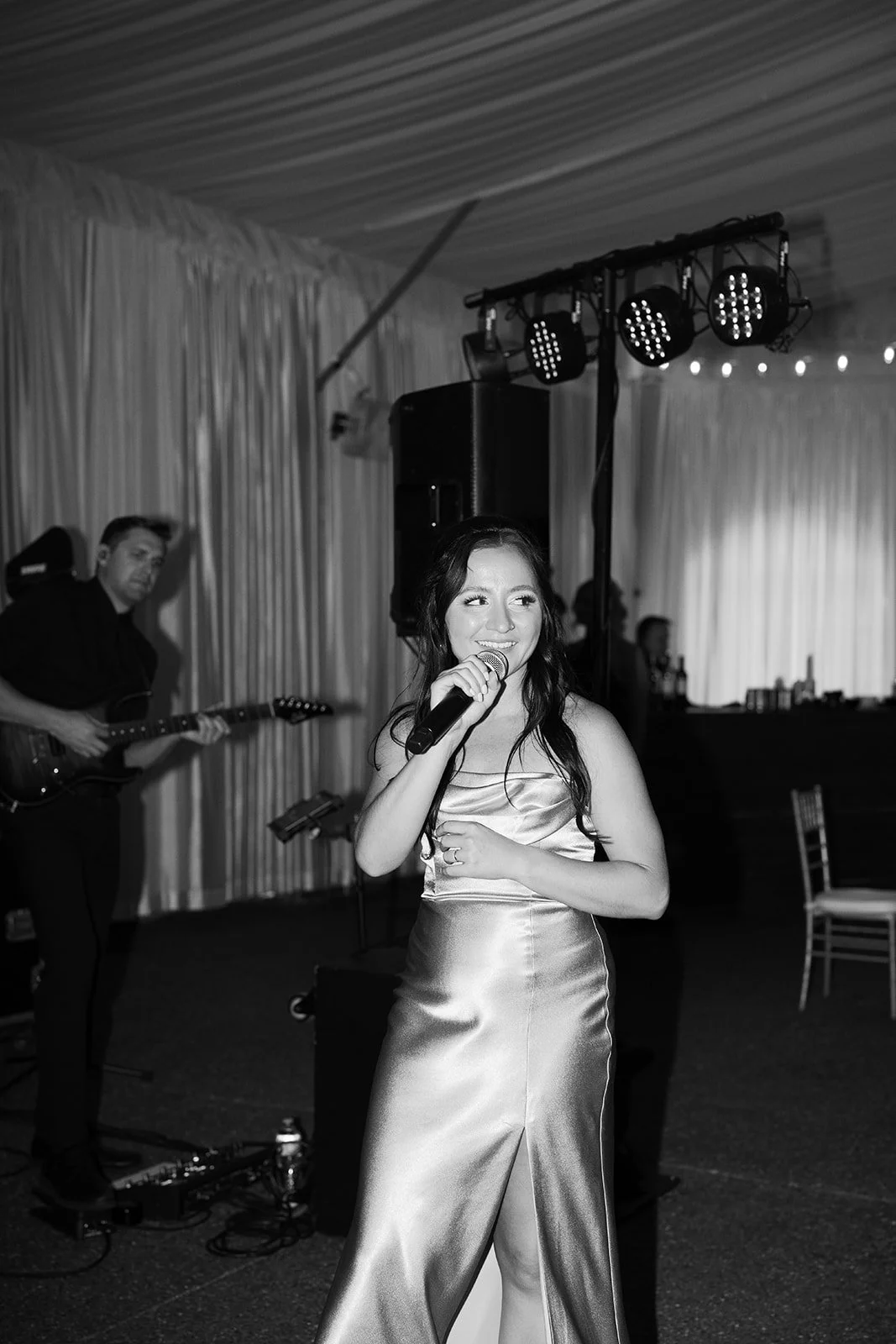 A woman singing into a microphone at a wedding reception or party, with a musician playing guitar in the background, in a decorated indoor venue.