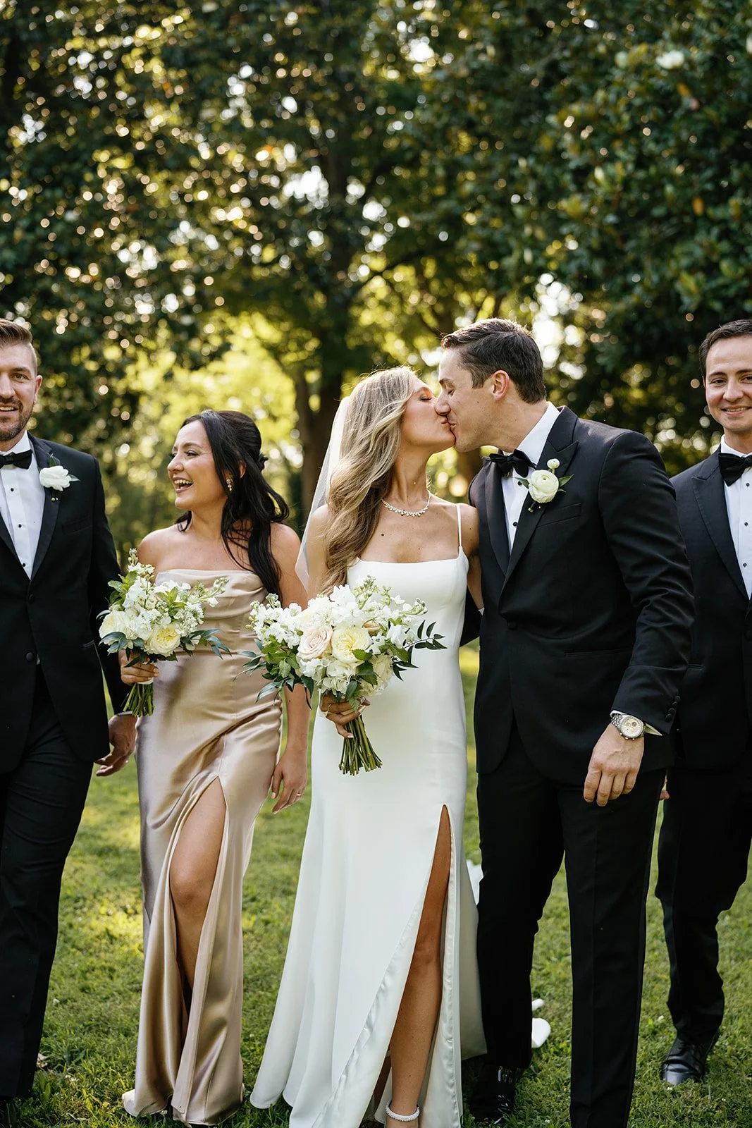 Bride and groom kiss surrounded by wedding party outdoors with trees in background.
