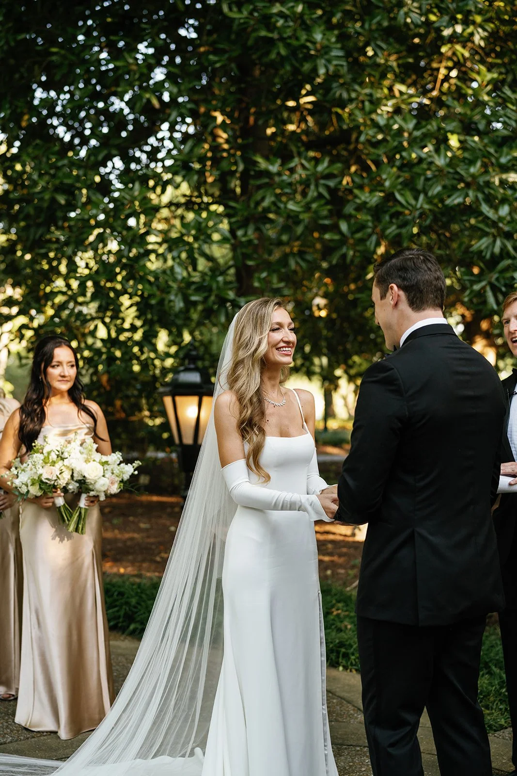 A bride and groom exchange vows during an outdoor wedding ceremony, with bridesmaids holding bouquets in the background.