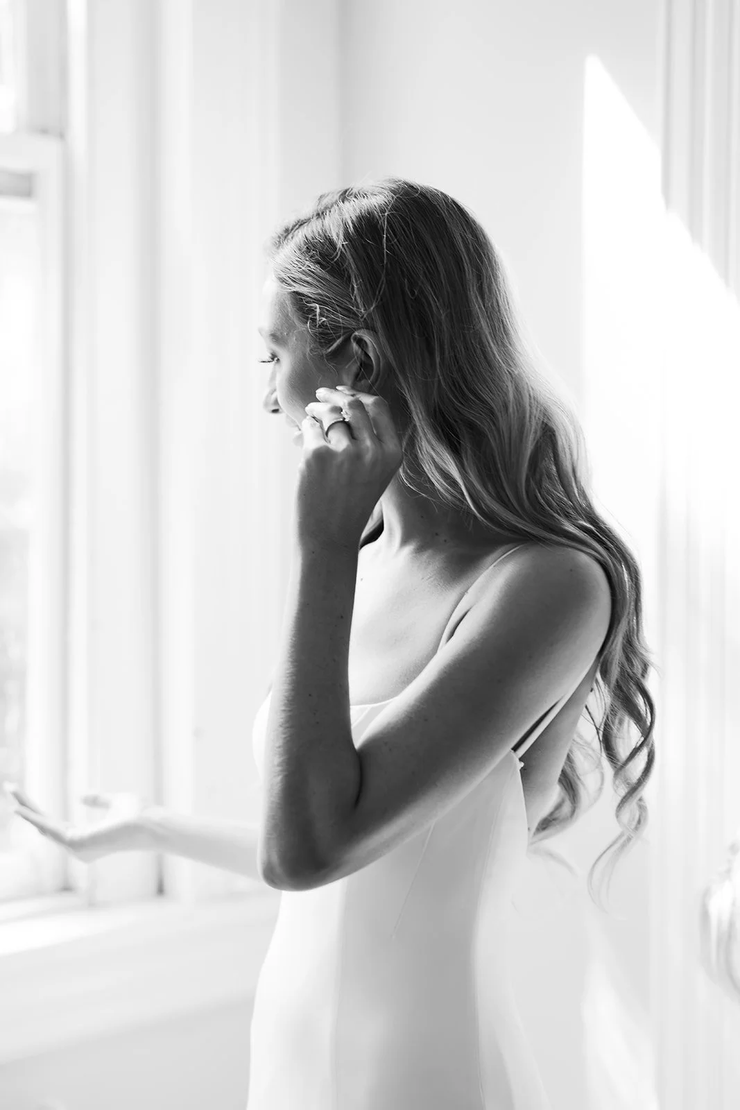 Black and white photo of a woman with long wavy hair looking to her right, touching her earring with her right hand, wearing a sleeveless dress, standing by a window.
