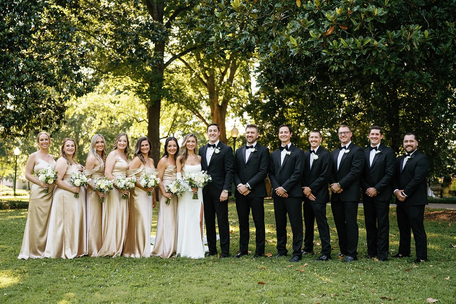 A wedding party of thirteen people standing outdoors on a grassy area with large trees in the background. The group includes bridesmaids in beige dresses holding bouquets and groomsmen in black tuxedos with bow ties. The bride and groom are in the ce