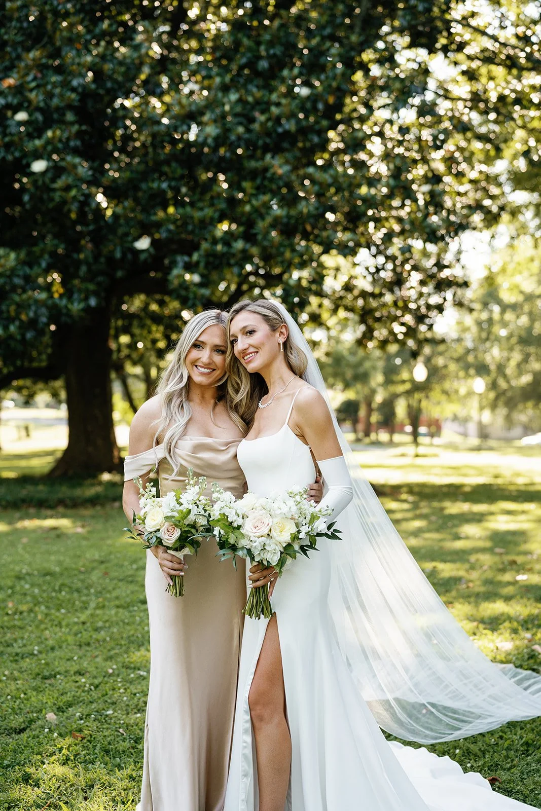 Two women in wedding attire holding bouquets and smiling outdoors in a park with large trees and sunlight.