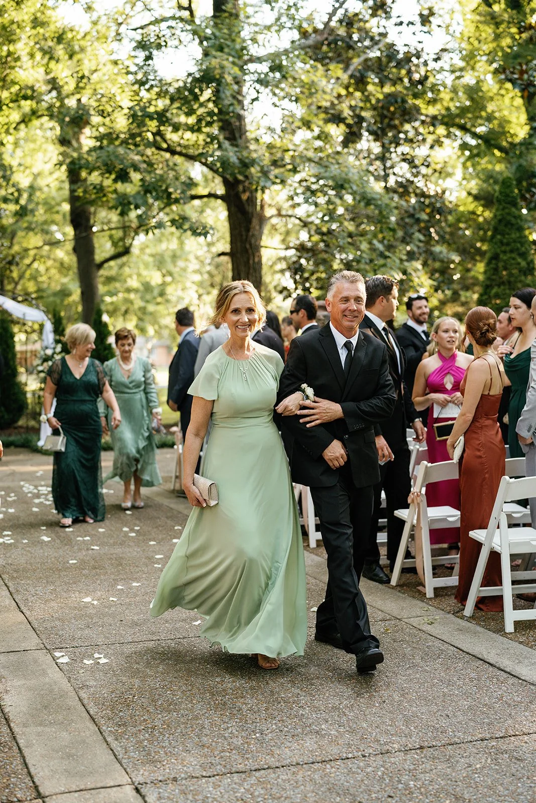 A woman in a light green dress and a man in a black suit walking arm in arm at an outdoor event, with other formally dressed guests in the background under trees.