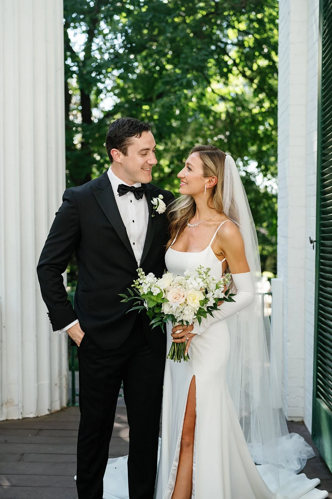 A newlywed couple on their wedding day outdoors. The groom is in a black tuxedo with a bow tie, and the bride wears a white wedding dress with a slit, holding a bouquet of white and blush flowers. They are smiling at each other against a background o