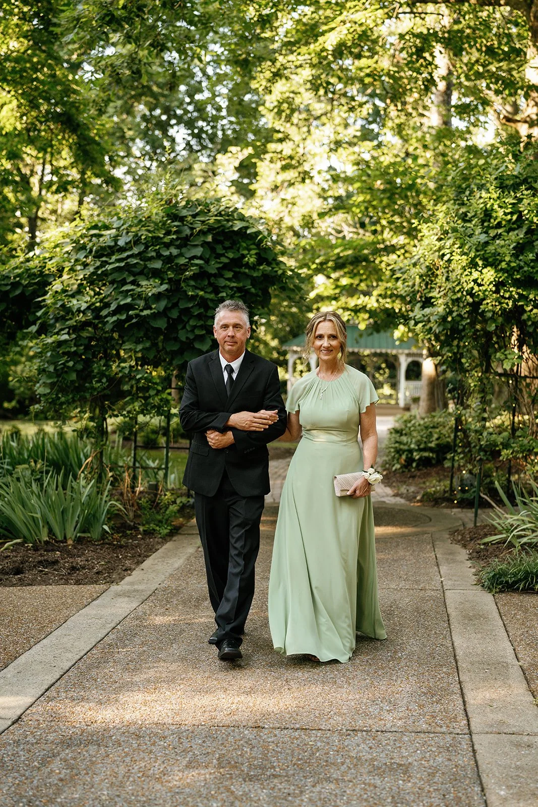 A man in a black suit and a woman in a light green gown walking arm in arm on a garden path surrounded by greenery.