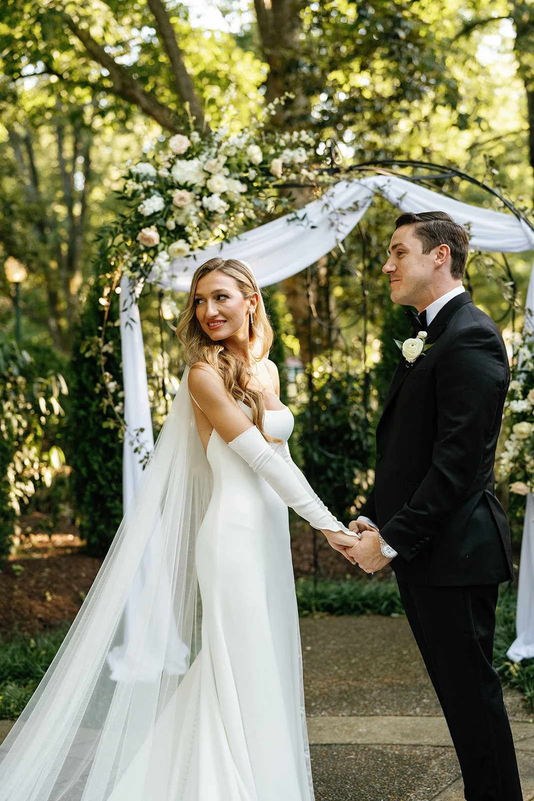 Bride and groom holding hands during outdoor wedding ceremony with floral arch and greenery.