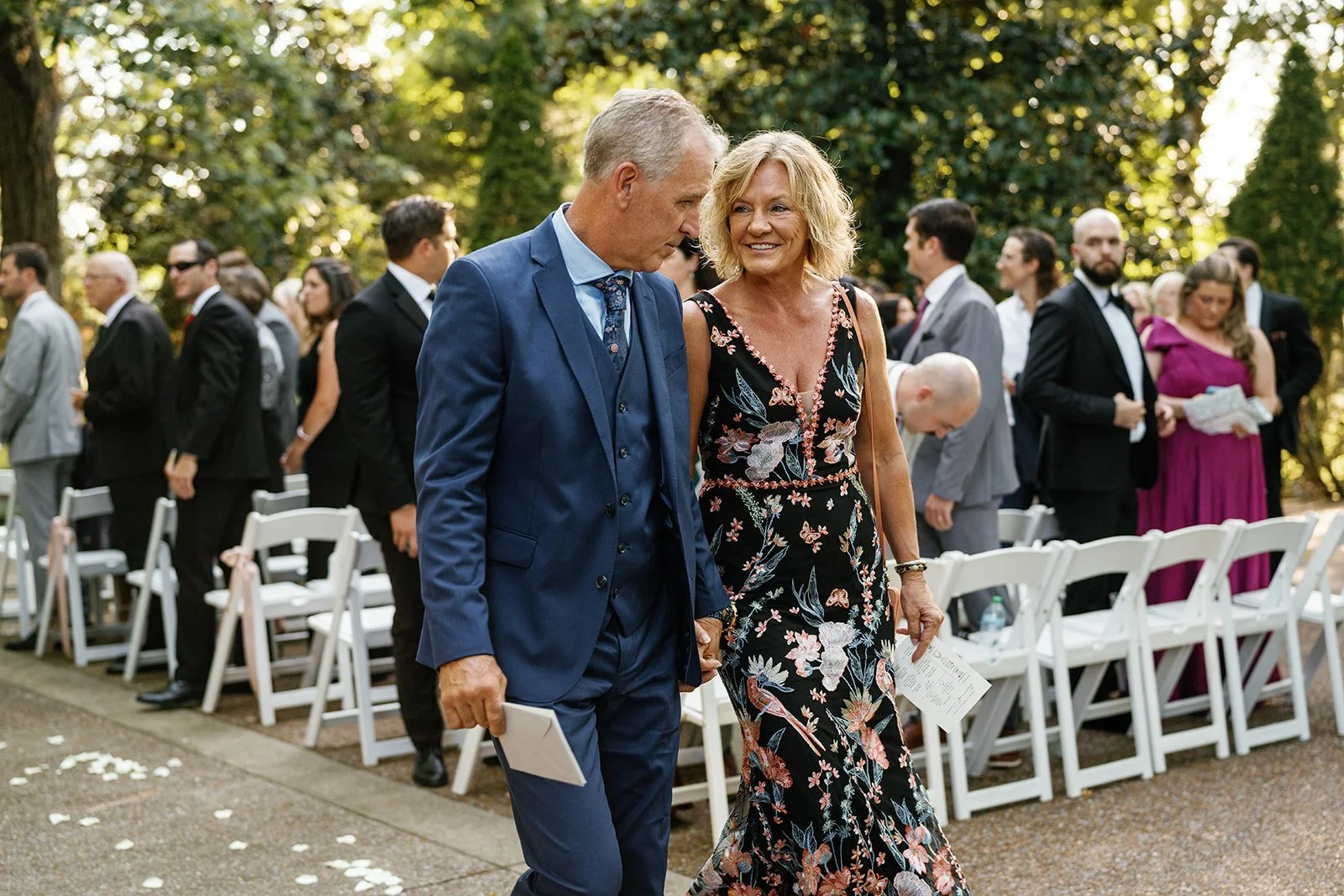 A man and woman walking hand in hand outdoors during a formal event, surrounded by seated and standing guests in formal attire, with trees and greenery in the background.