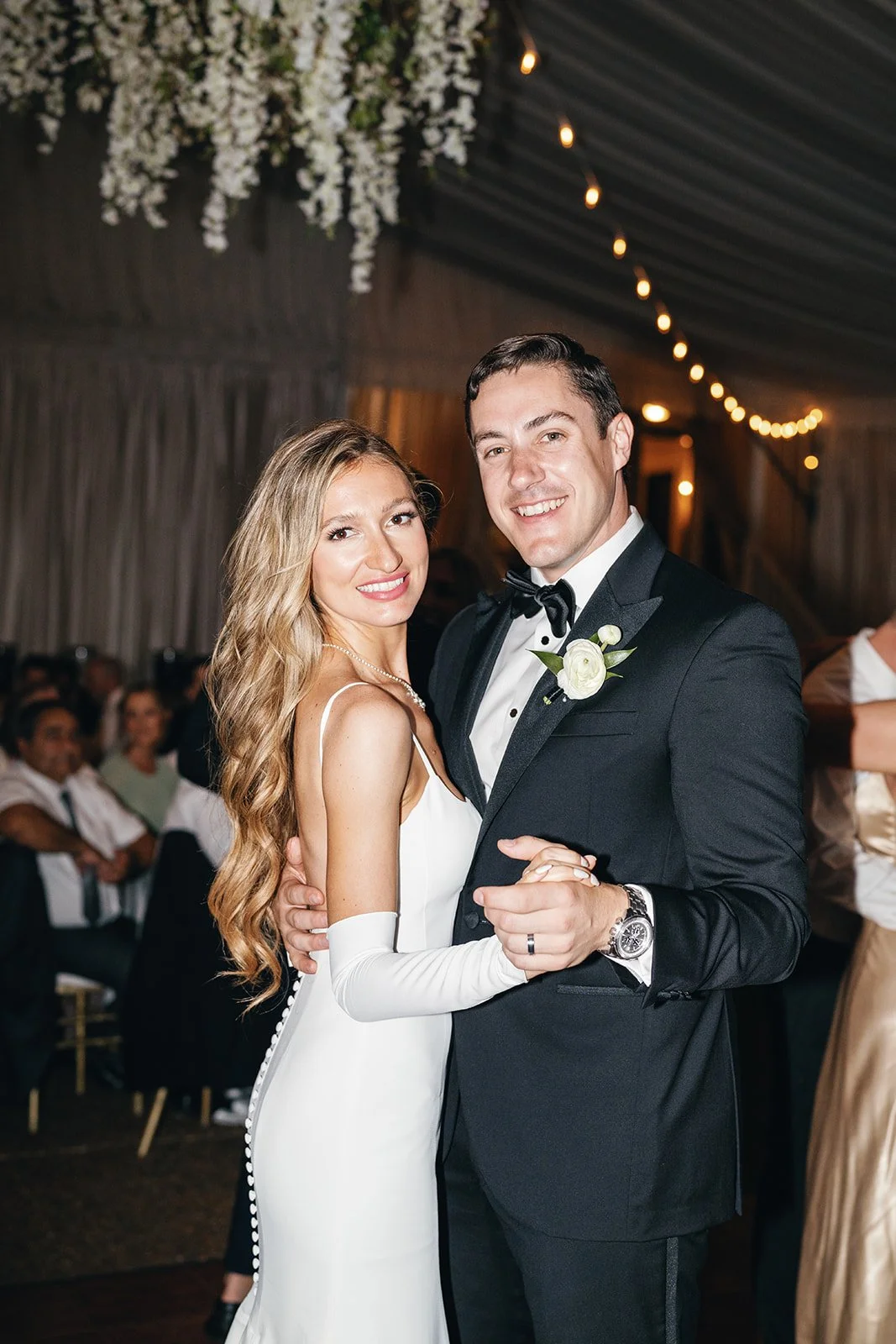A bride and groom dancing at their wedding reception inside a decorated venue with string lights and floral arrangements.