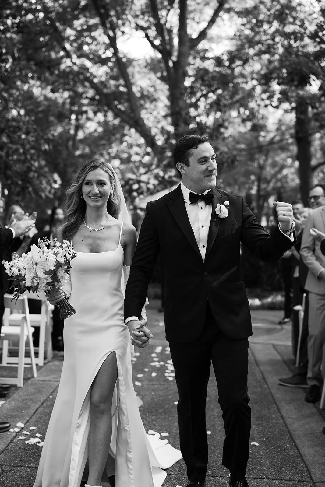 Black and white photo of a newlywed couple holding hands and walking outdoors. The bride is in a white dress with a slit, holding a bouquet and smiling. The groom is in a tuxedo with a bow tie, looking happy and raising his fist.