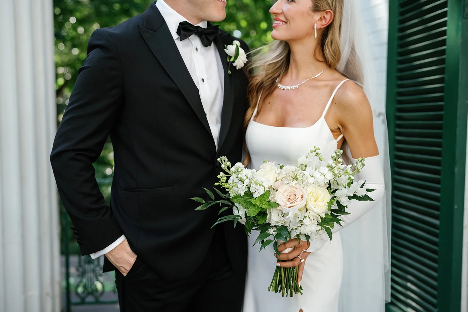 A bride and groom on their wedding day, with the bride holding a bouquet of white and blush flowers, standing near white columns and green shutters.