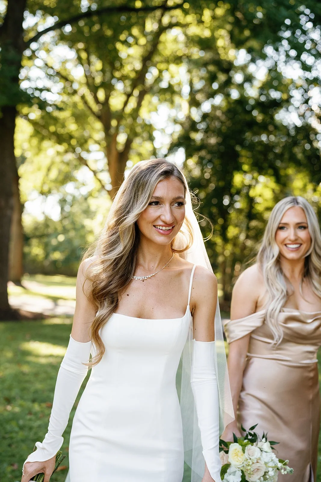 A bride in a white wedding gown with long gloves and a necklace standing outdoors, smiling, with a bridesmaid in a beige dress holding a bouquet visible in the background.