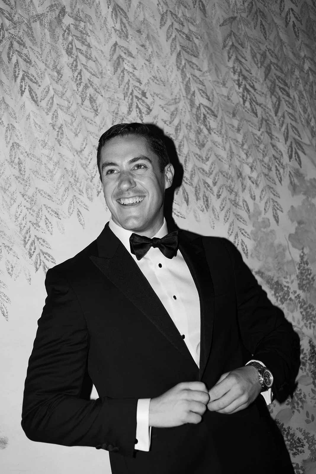 A young man in a tuxedo with a bow tie smiling and adjusting his jacket, standing in front of a patterned wall.