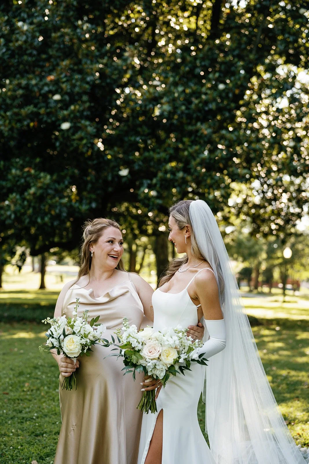 A bride and woman in a park, smiling and holding bouquets of white flowers.