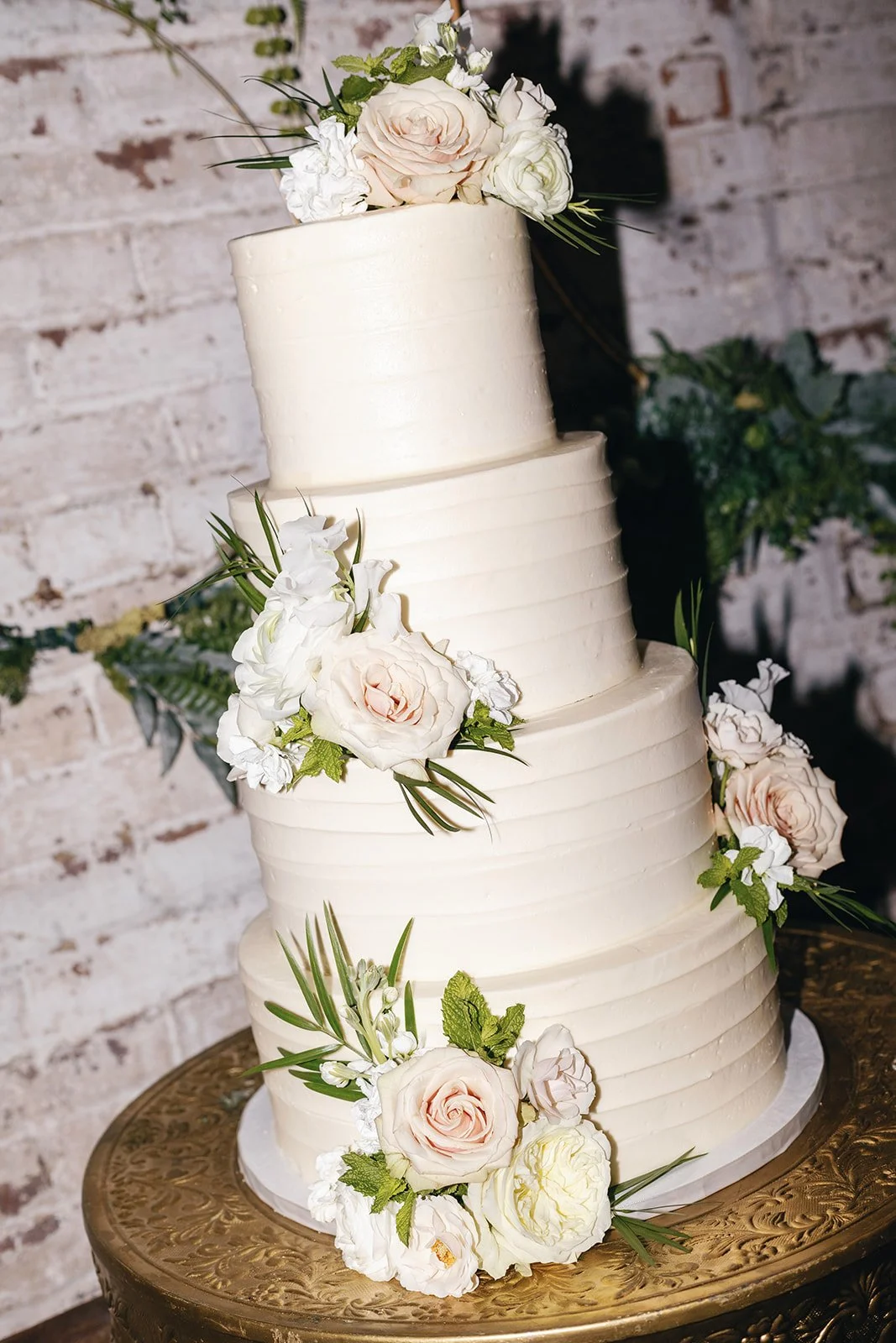 Three-tiered wedding cake decorated with white and blush pink roses, white peonies, and greenery, placed on an ornate gold stand against a white brick wall.