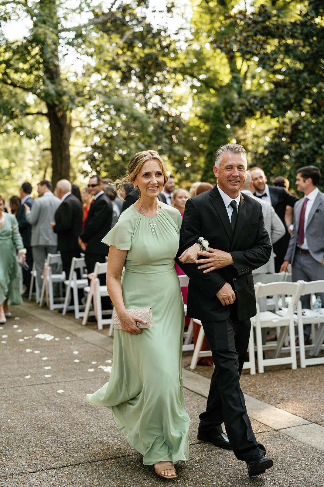 A woman in a long, light green dress and a man in a black tuxedo walk arm-in-arm at an outdoor wedding ceremony, with several guests seated and standing in the background among trees.