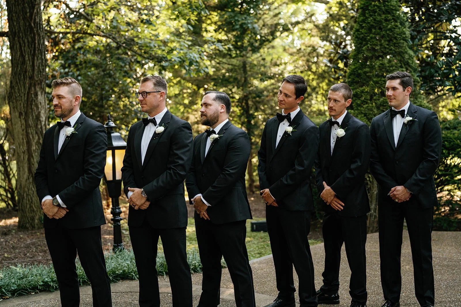 Six men dressed in black tuxedos with white shirts and black bow ties, standing outdoors on a pathway in a garden, during daylight. They are lined up, facing slightly to the right, with trees and greenery in the background.