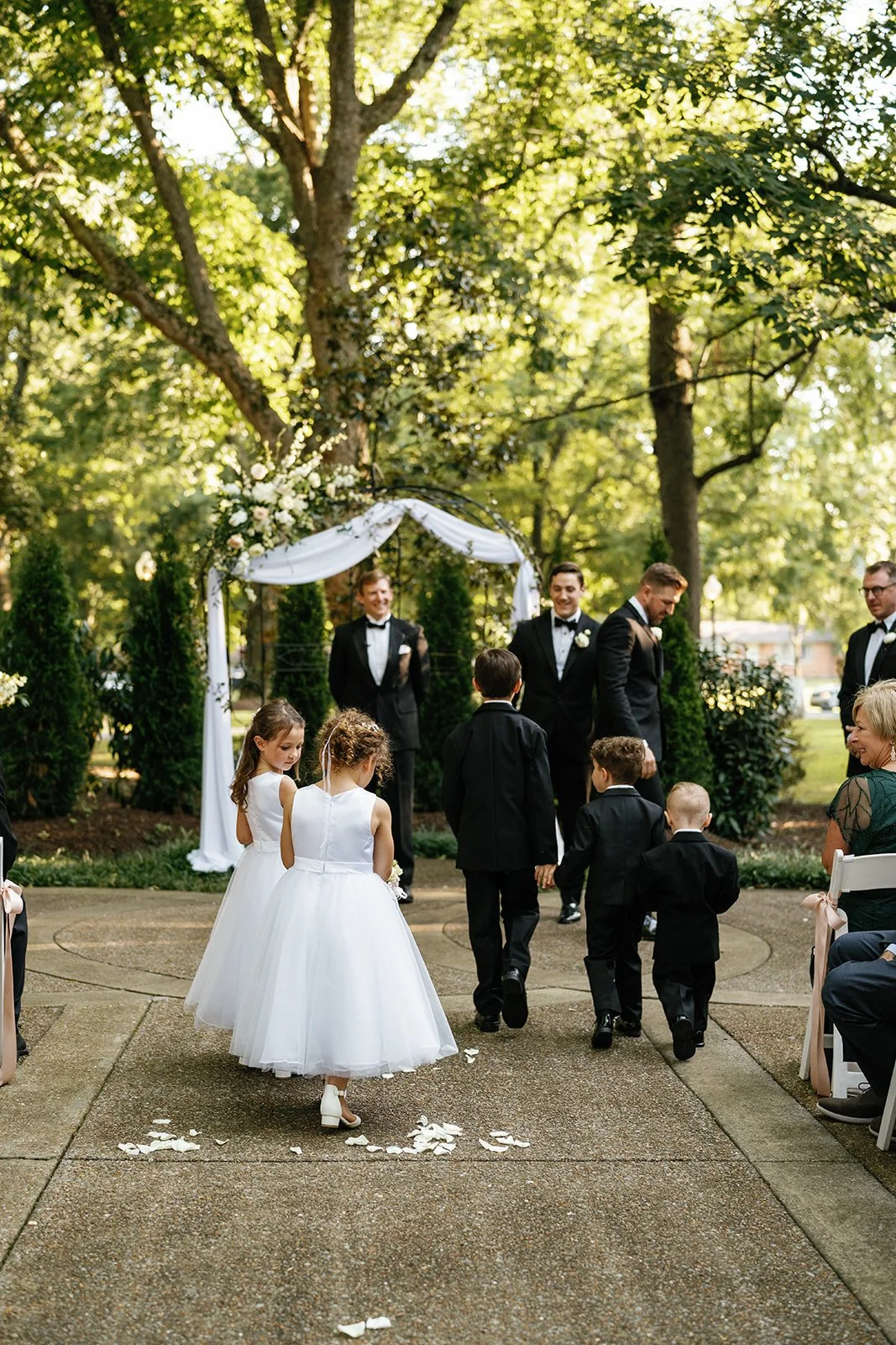 A wedding ceremony taking place outdoors under a large tree with green leaves. Children in white dresses and black suits walk towards the altar, which is decorated with white fabric and flowers. Guests are seated on either side, watching the processi