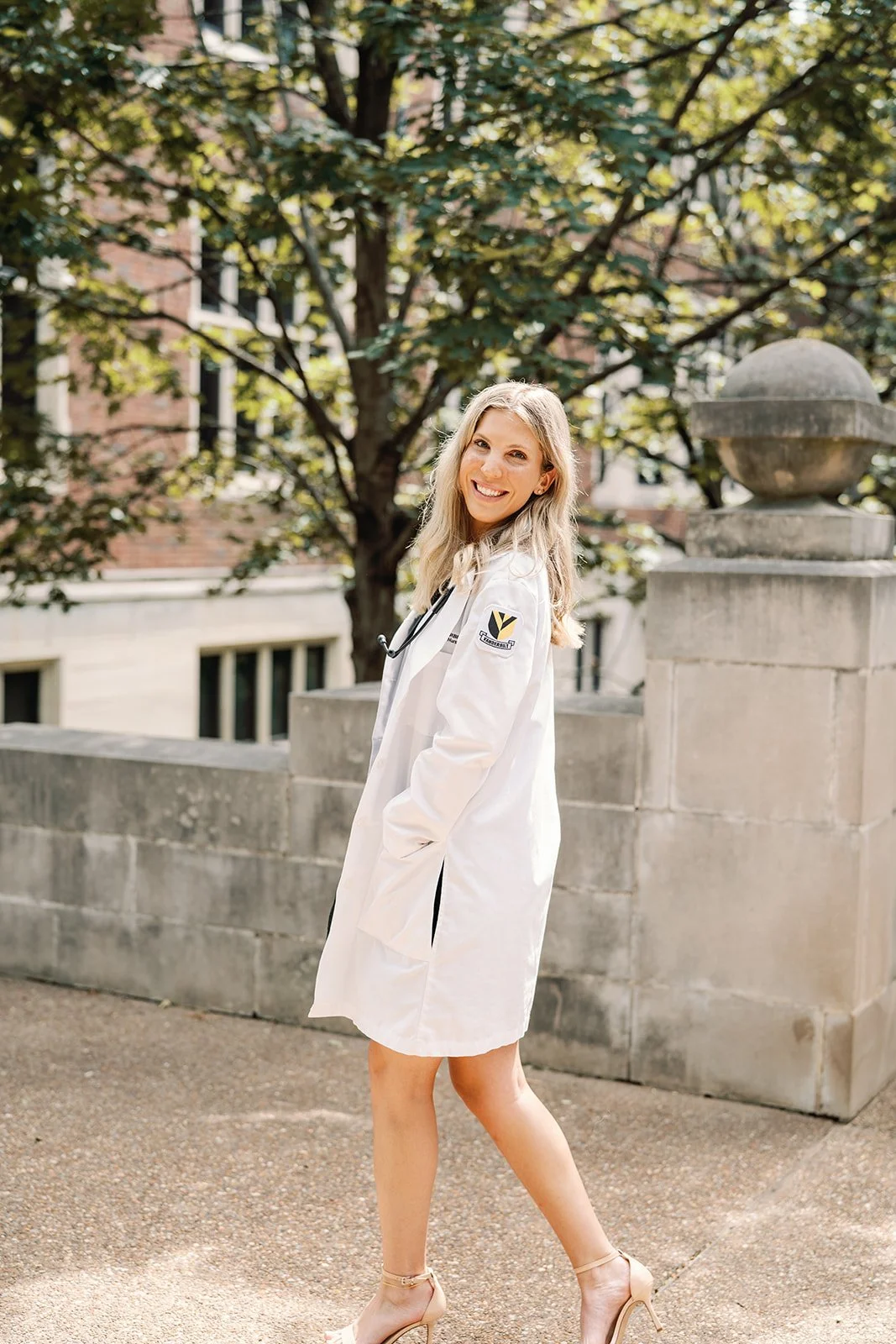 A Vanderbilt grad student, standing outdoors near a stone wall with a tree and brick building in the background, smiling at the camera.