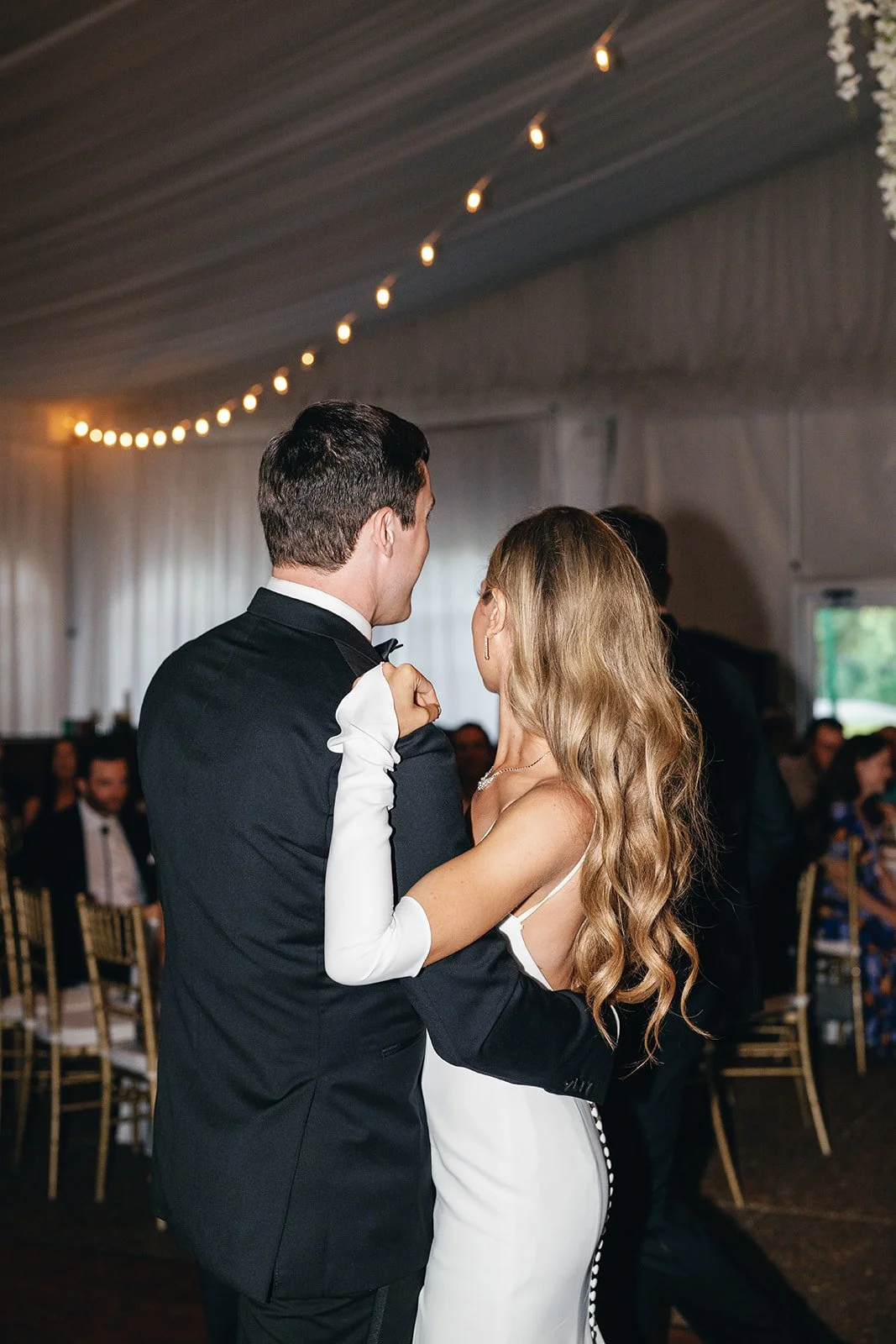 A bride and groom dance at their wedding reception inside a decorated hall with string lights.