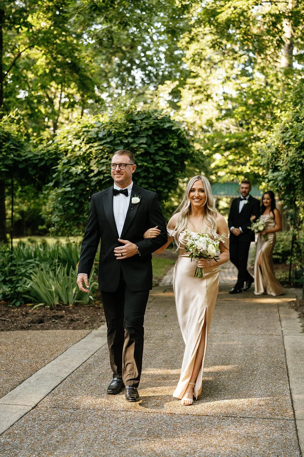 A bride with long blonde hair in a champagne-colored gown holding a bouquet, walking arm-in-arm with a man in a black tuxedo, with two more people in formal attire walking behind them in a lush garden setting.