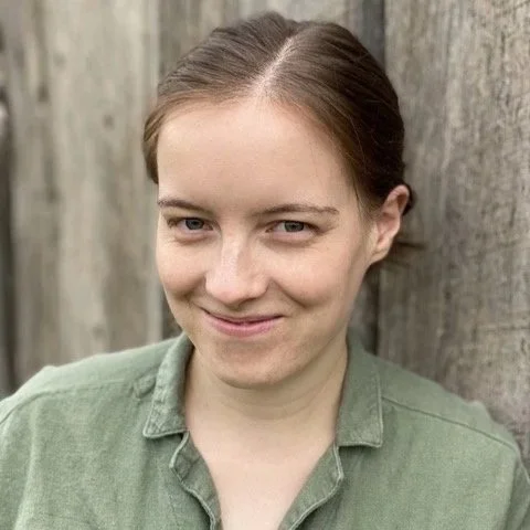 Portrait of a young woman with light brown hair in a ponytail, wearing a green shirt, standing outdoors against a wooden fence.