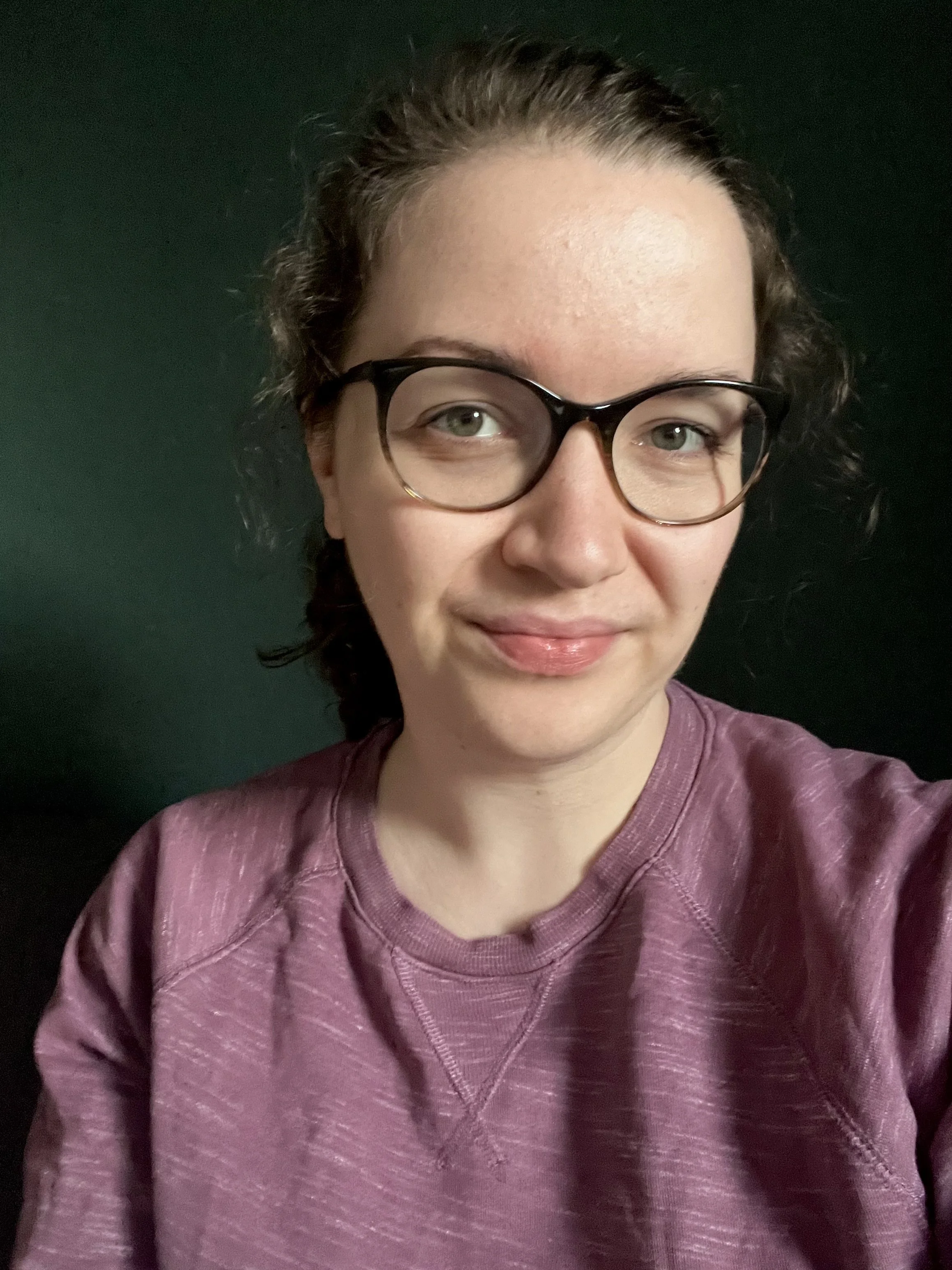 A woman with glasses and dark curly hair wearing a purple sweatshirt, smiling slightly, against a dark green background.