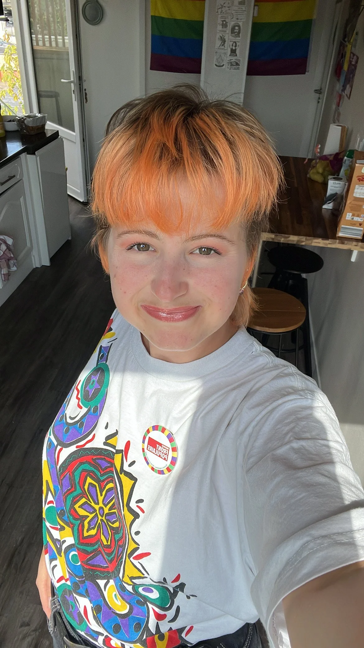 A young person with shoulder-length orange hair, smiling and taking a selfie in a kitchen. They are wearing a white t-shirt with colorful abstract artwork and a round sticker.