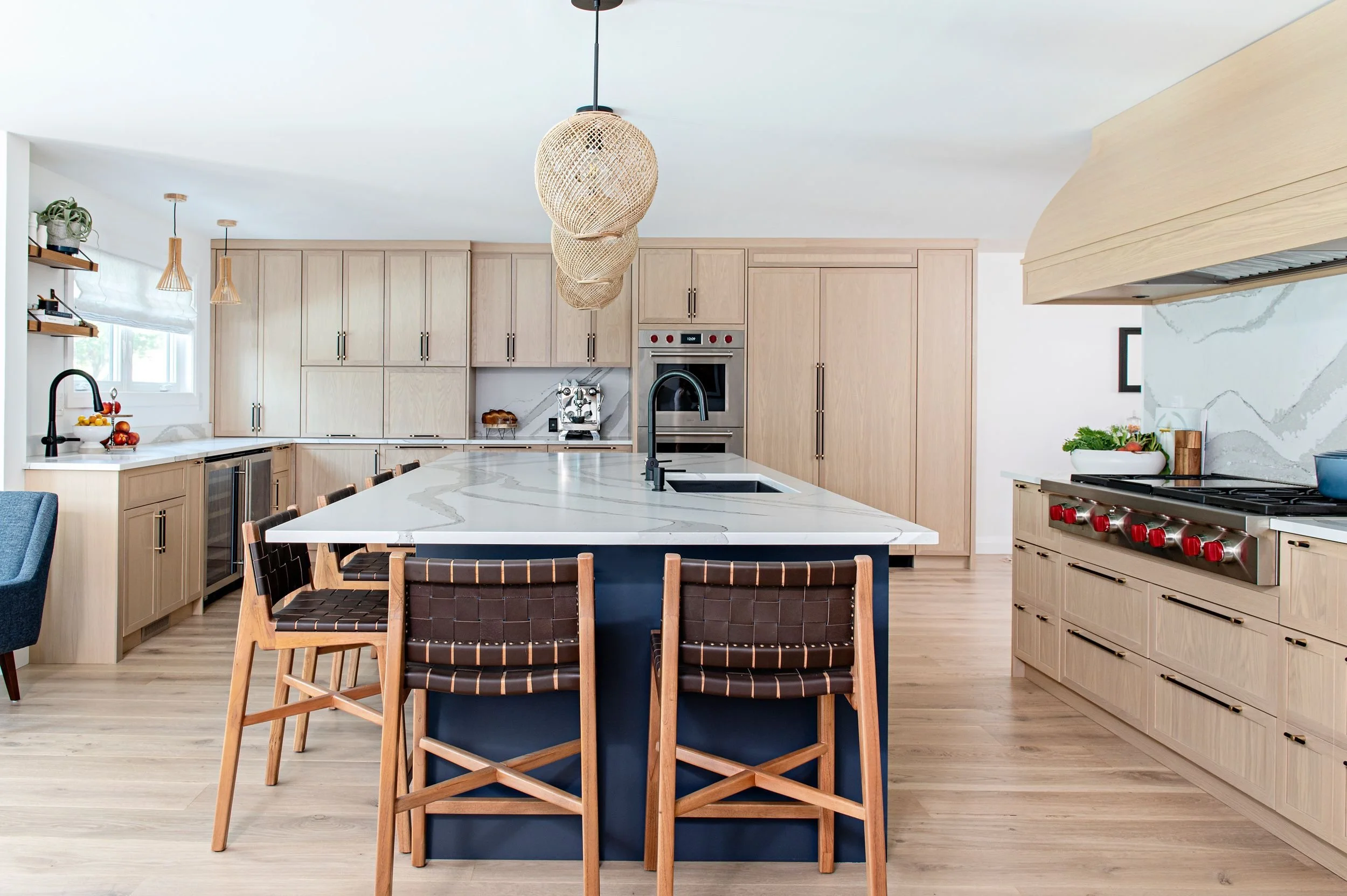 Modern kitchen with light wooden cabinets, white marble countertops, and a large island with seating. Black fixtures, a gas stove with red knobs, and a wooden range hood. Light hardwood floors, decorative pendant lights, and potted plants.