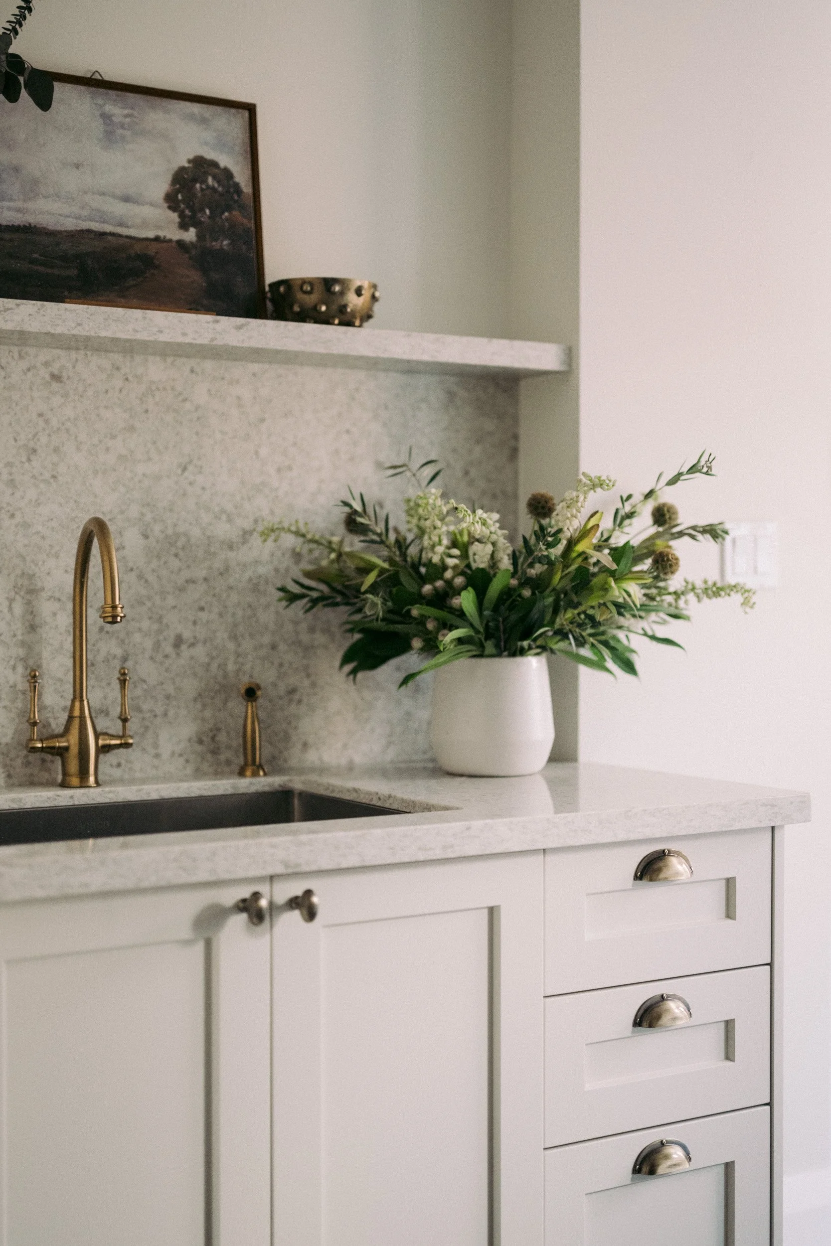 A kitchen countertop with a white vase of green and white flowers, a gold faucet, and a cabinet with drawers.