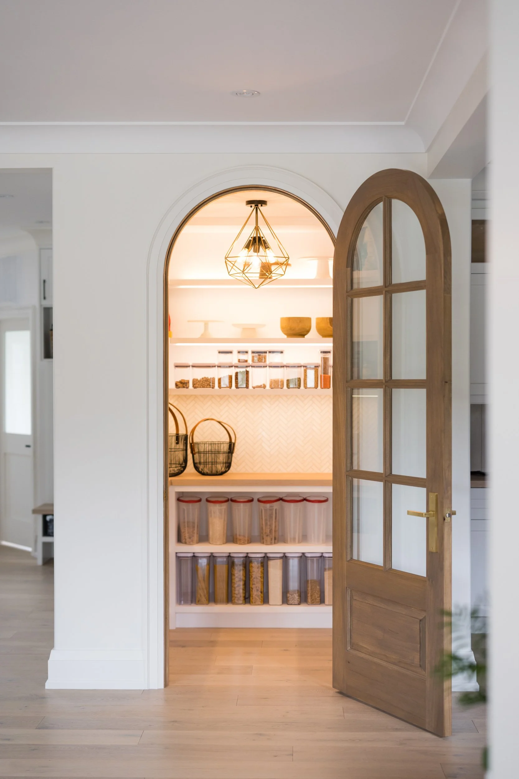Open wooden door leading into a pantry with shelves filled with containers of dry goods, bowls, and baskets, illuminated by warm lighting