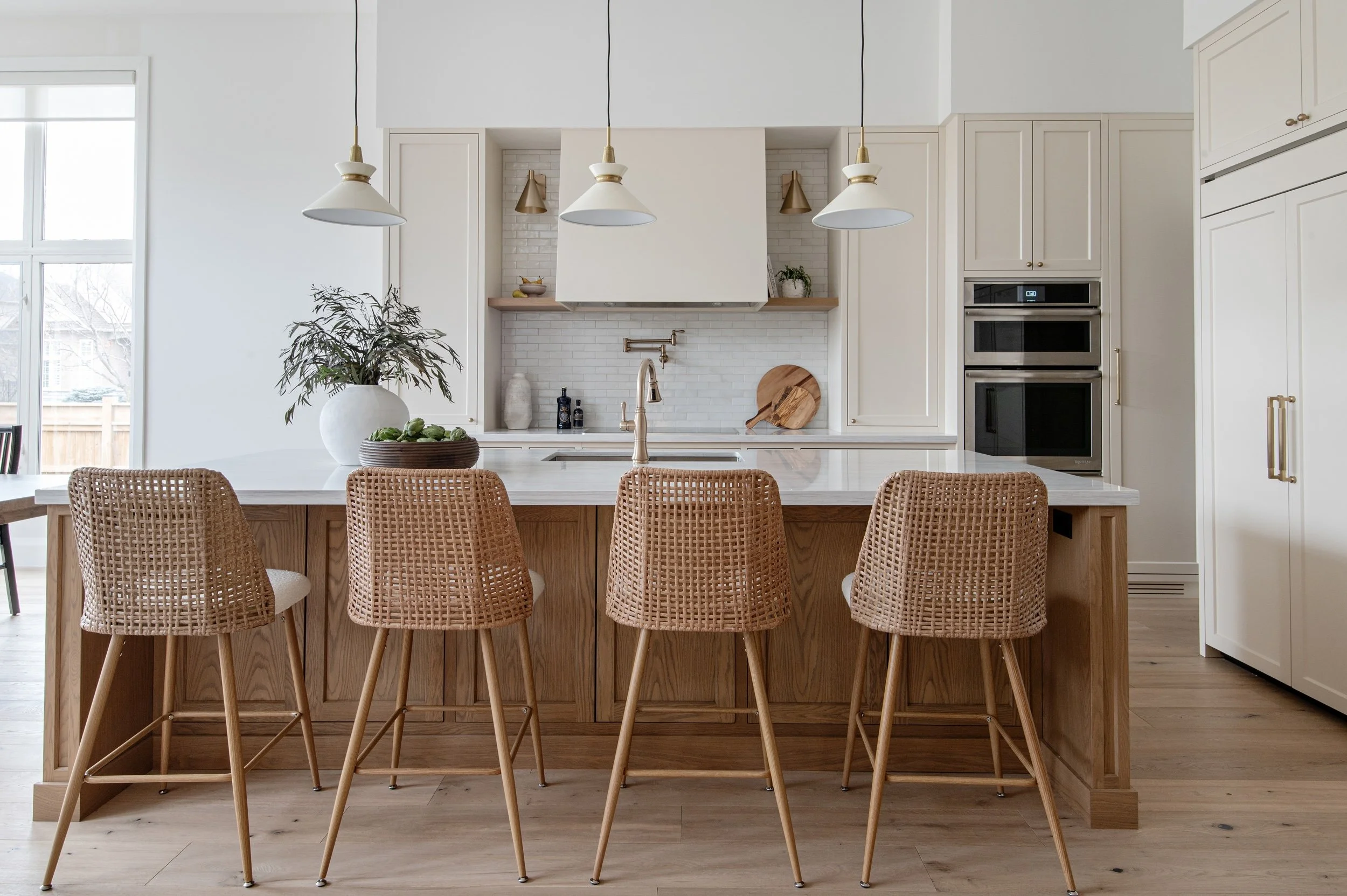 Modern kitchen with white cabinetry, wooden island with four woven rattan barstools, white countertops, pendant lights, and built-in double ovens.