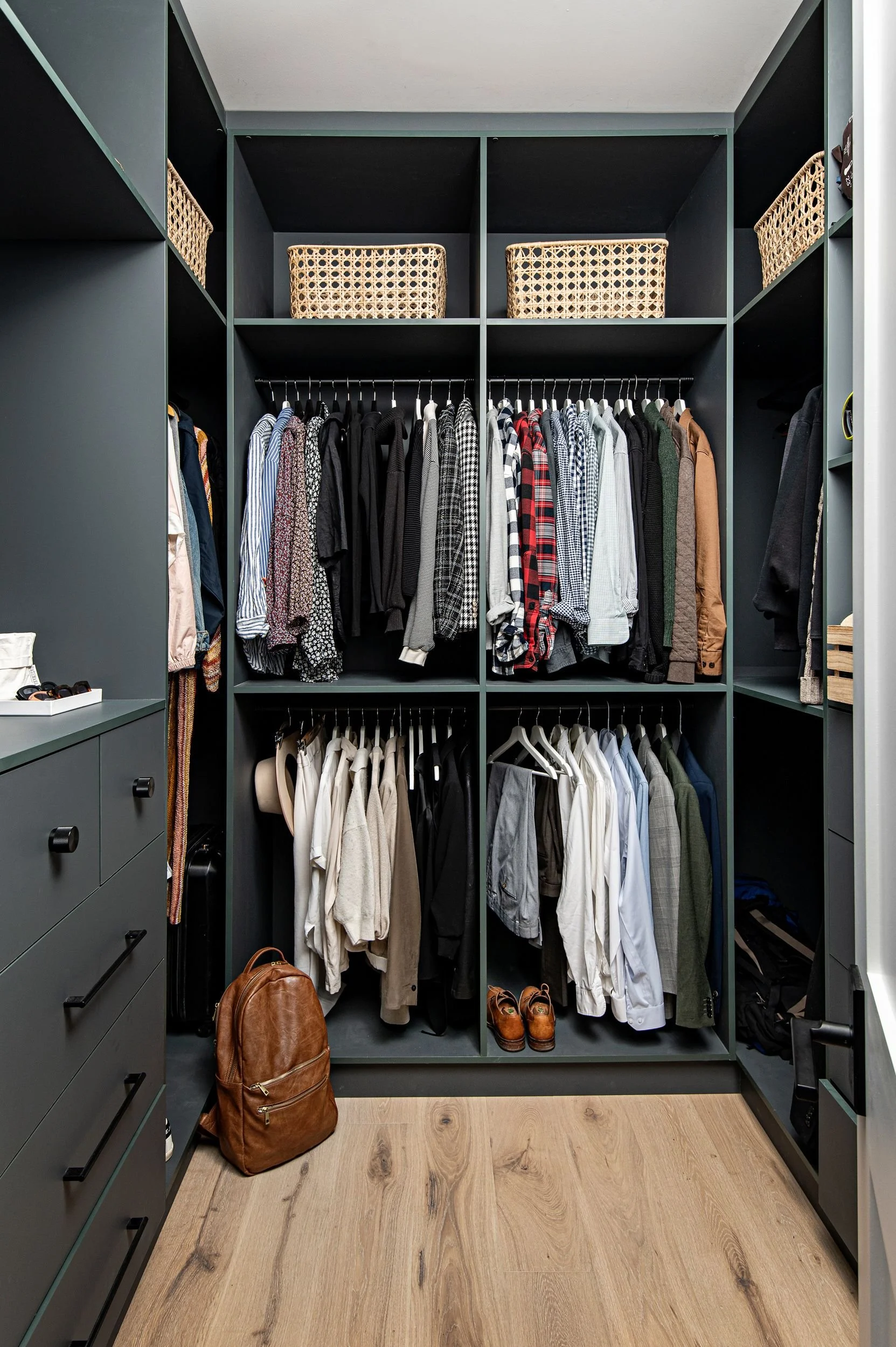 A organized walk-in closet with dark green shelving holding clothes, baskets, and accessories. There are clothes hanging on rods, shoes on the bottom, and a brown backpack on the floor.