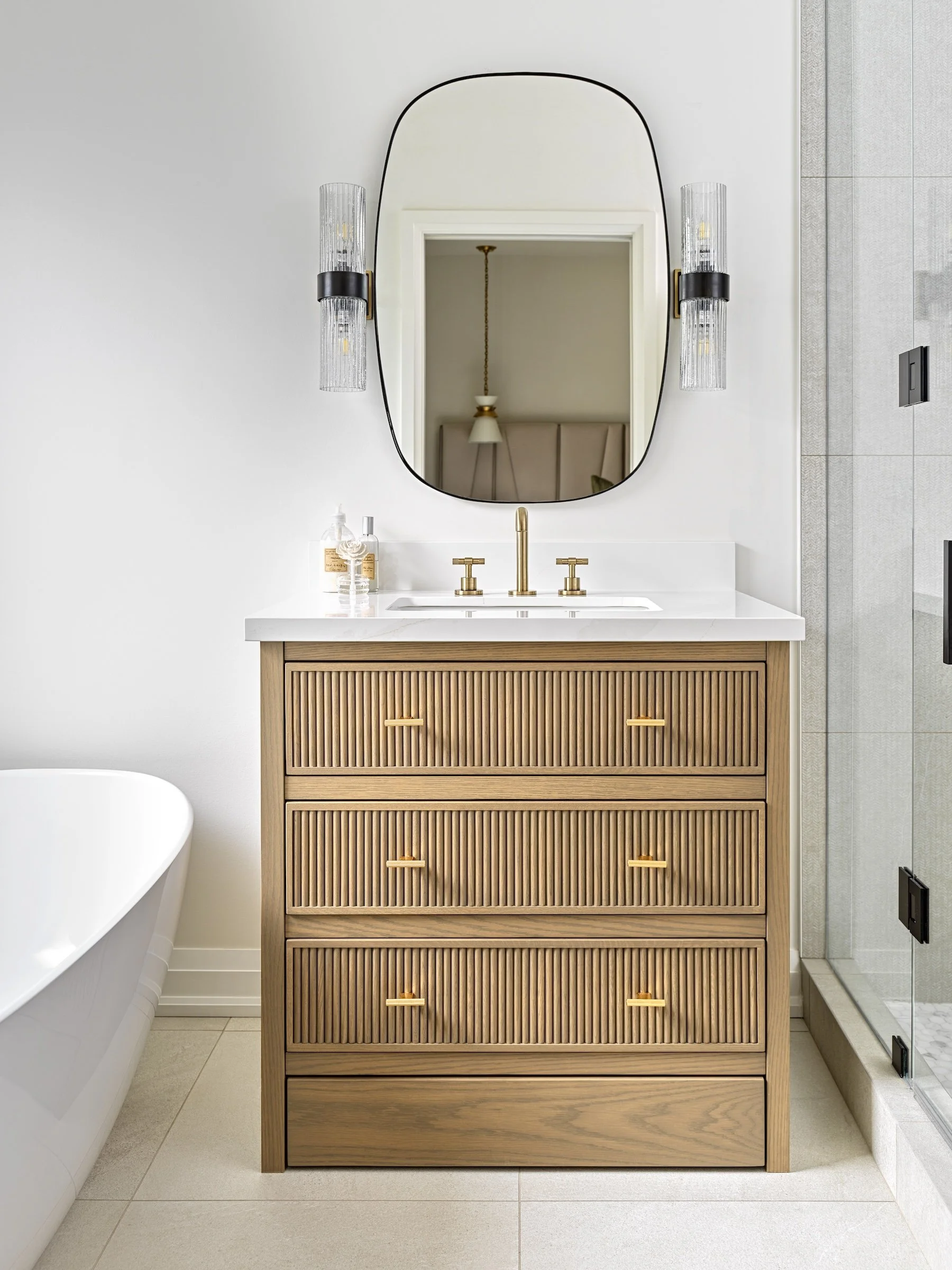 Modern bathroom with wooden vanity, white countertop, bronze faucet, black-framed mirror, wall-mounted light fixtures, partial shower enclosure, bathtub.
