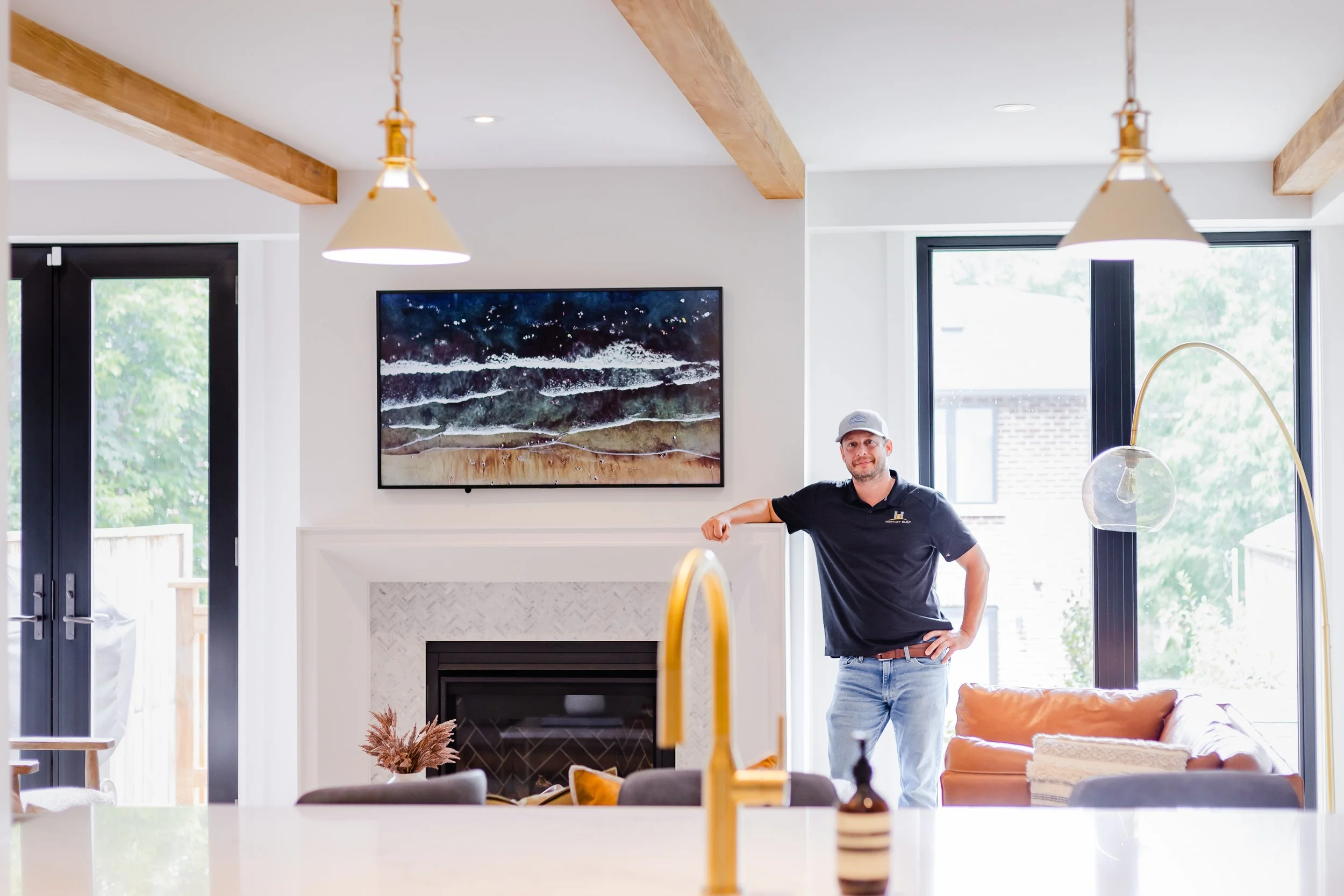 A man wearing a black polo shirt, light blue jeans, and a gray cap standing indoors near a fireplace, with his right arm resting on the fireplace mantle. Modern living room with large windows, a sofa, wall-mounted artwork, and a kitchen island with a golden faucet in the foreground.