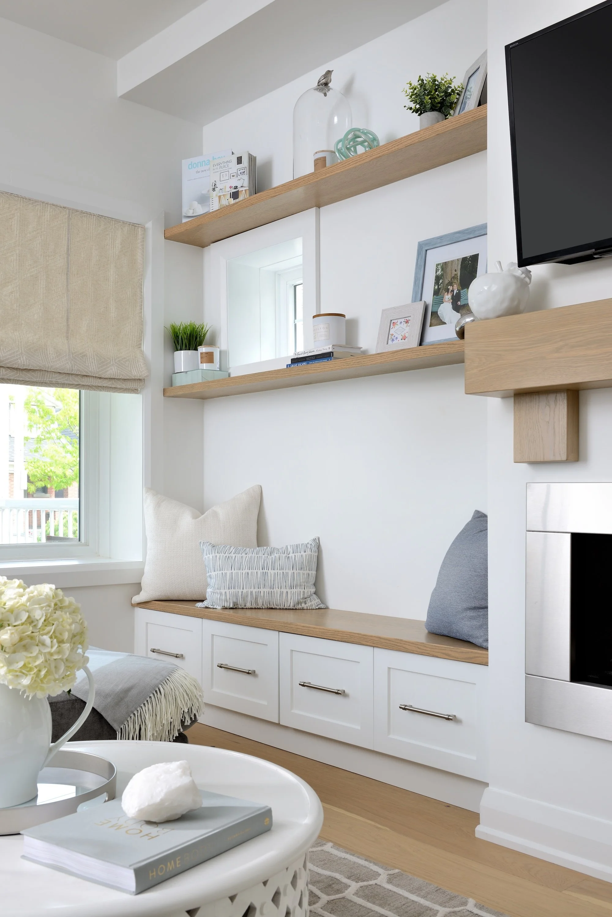 Cozy living room corner with window seat, cushions, built-in storage, floating shelves with decorative items, and a wall-mounted flat-screen TV.