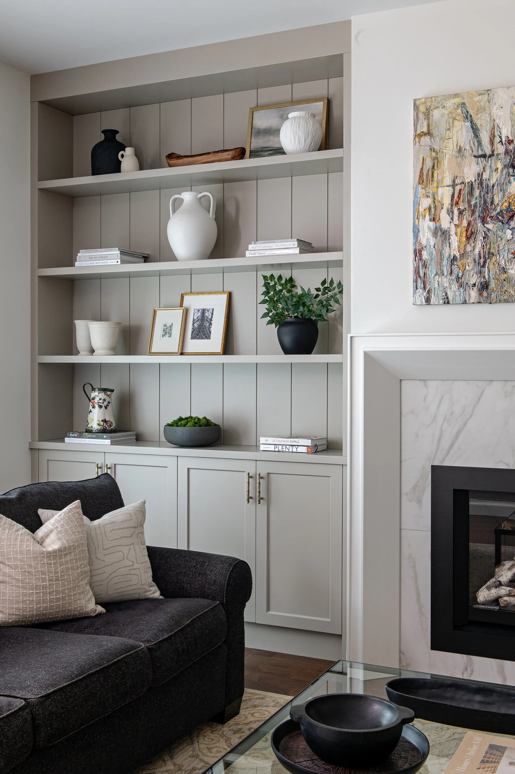 A living room with a built-in bookshelf filled with decorative vases, framed photos, books, and a plant. A black sofa with light-colored pillows is in the foreground, next to a glass coffee table with black bowls.