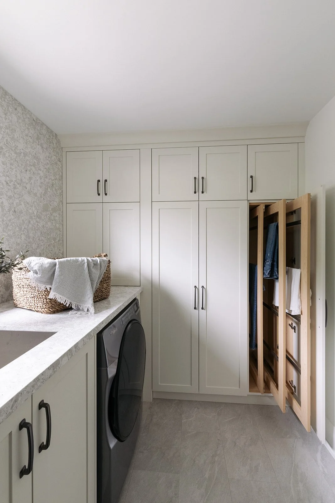 Laundry room with white cabinets, a washing machine, a basket with a towel, and a wooden clothes drying rack with hanging clothes.