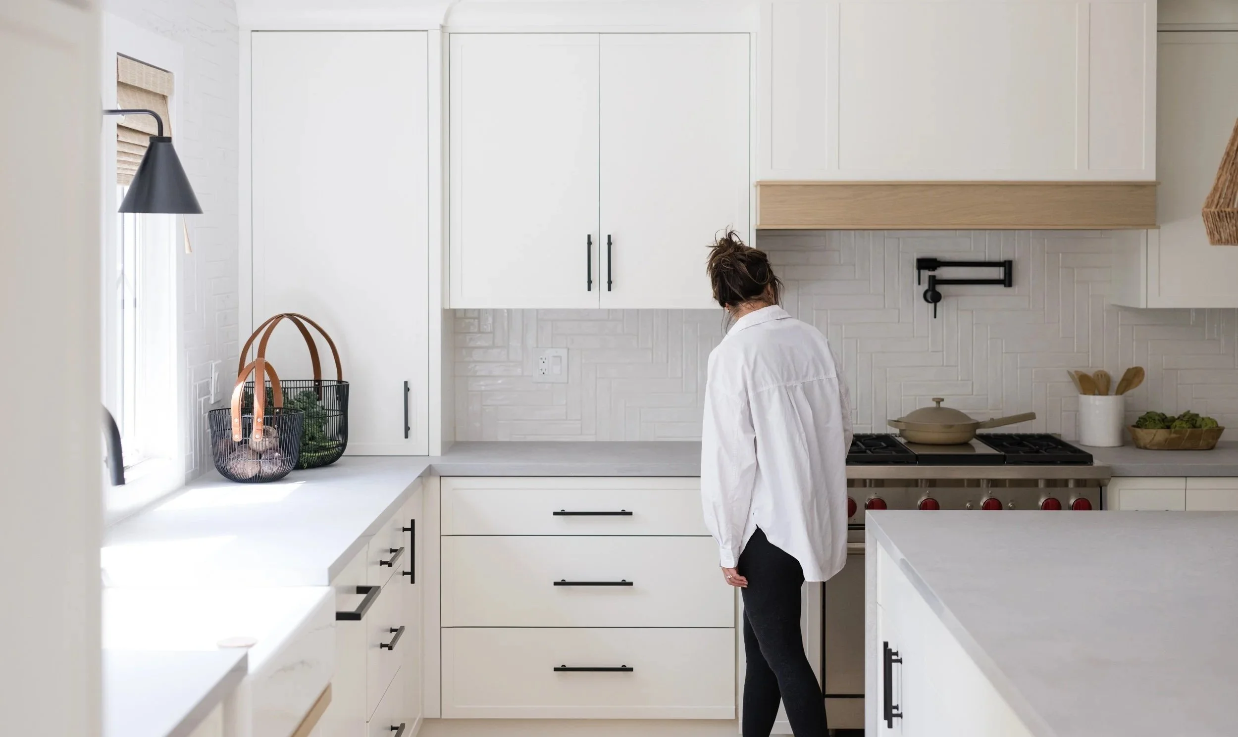 A woman standing near a stove in a modern white kitchen with baskets of vegetables on the counter.