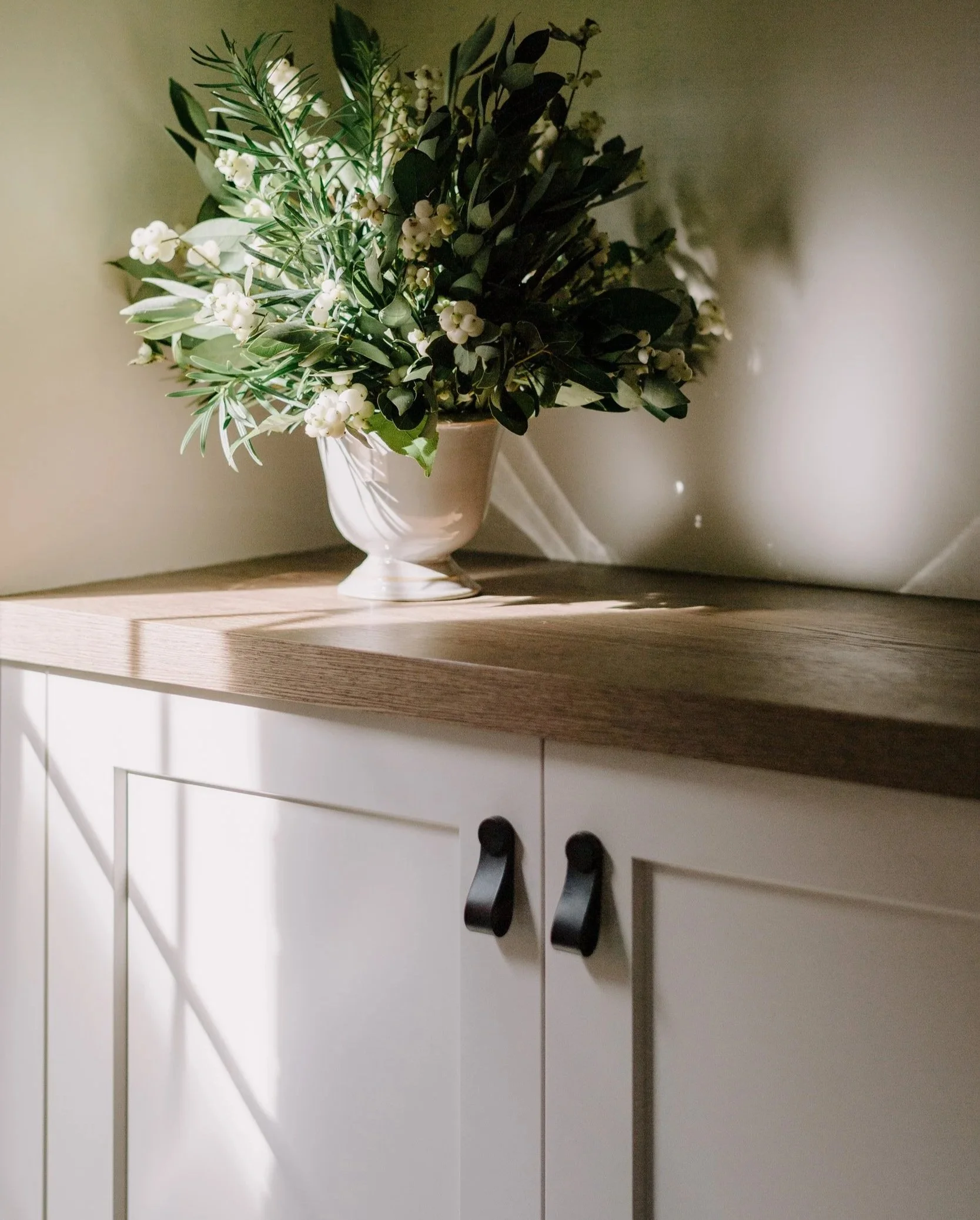A bouquet of white flowers and green foliage in a white vase on a wooden cabinet with sunlight casting shadows.