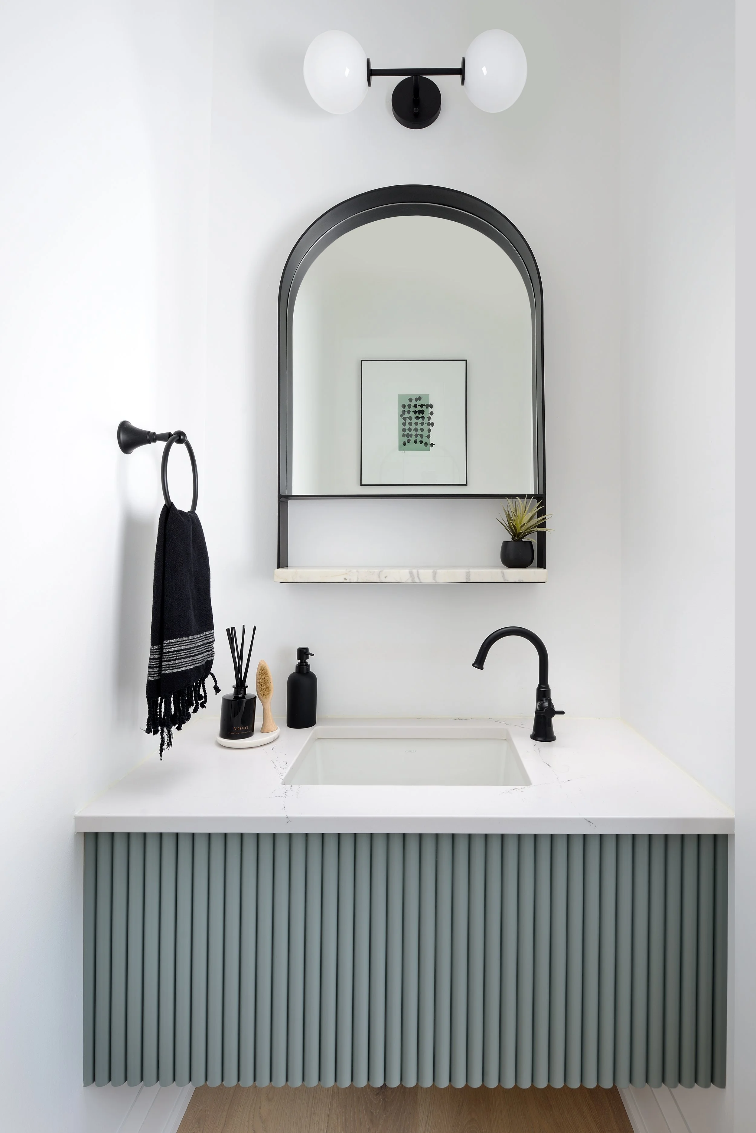 Modern bathroom vanity with white sink, black faucet, black mirror, wall-mounted light fixture with two globes, black towel ring with a black towel, black soap dispenser, reed diffuser, small black planter with a plant, and framed artwork reflected in the mirror.