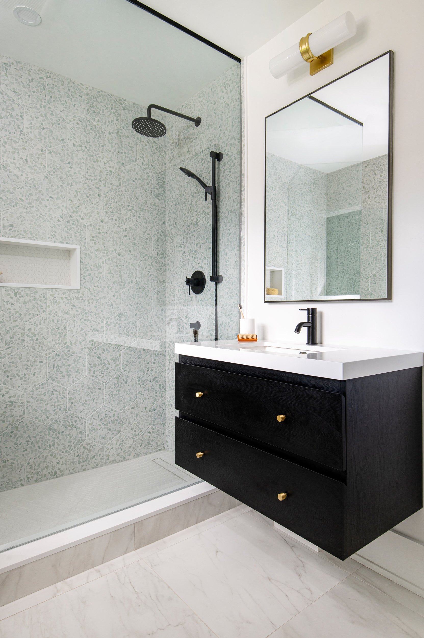 Modern bathroom with a black vanity, white countertop, large mirror, and a glass shower with black fixtures and gray mosaic tiles.