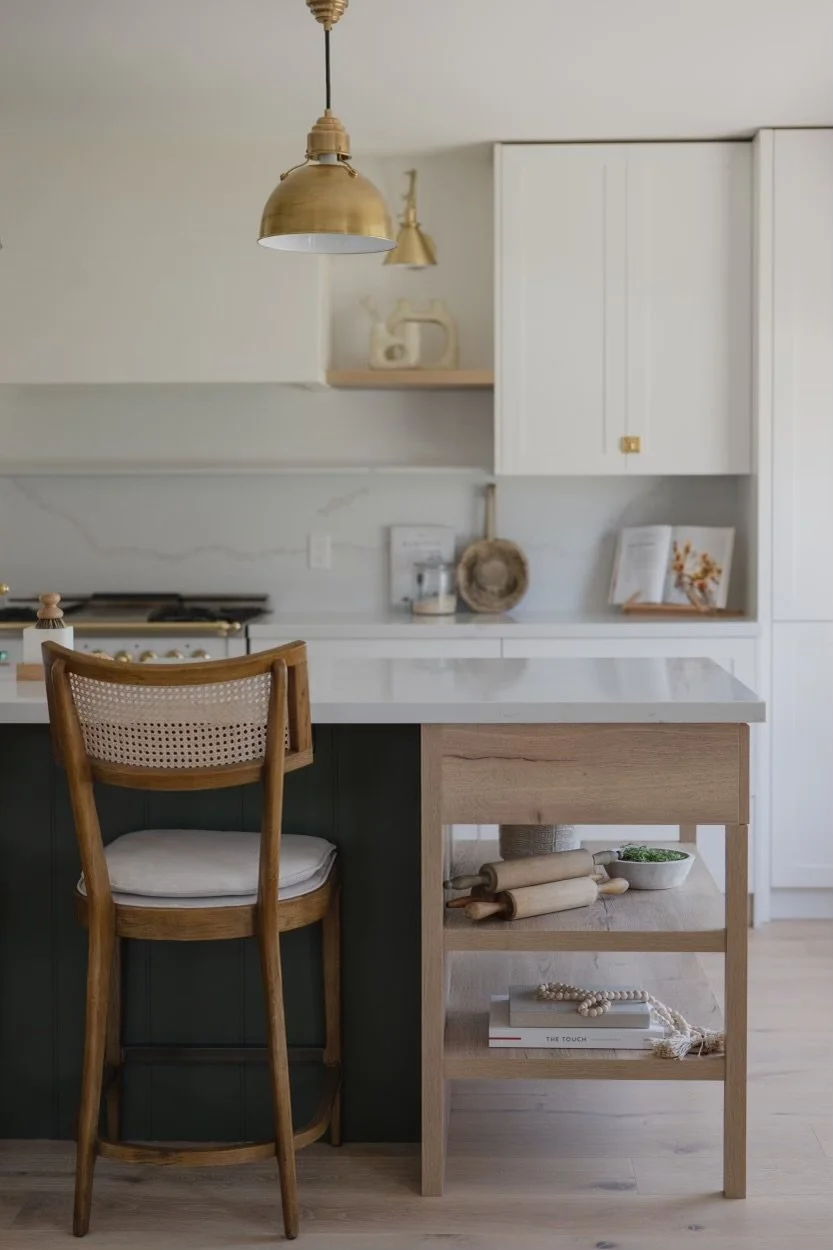 Modern kitchen with white cabinets, a marble countertop, hanging gold pendant lights, a wooden chair, a small rolling cart with kitchen items, and decor items.