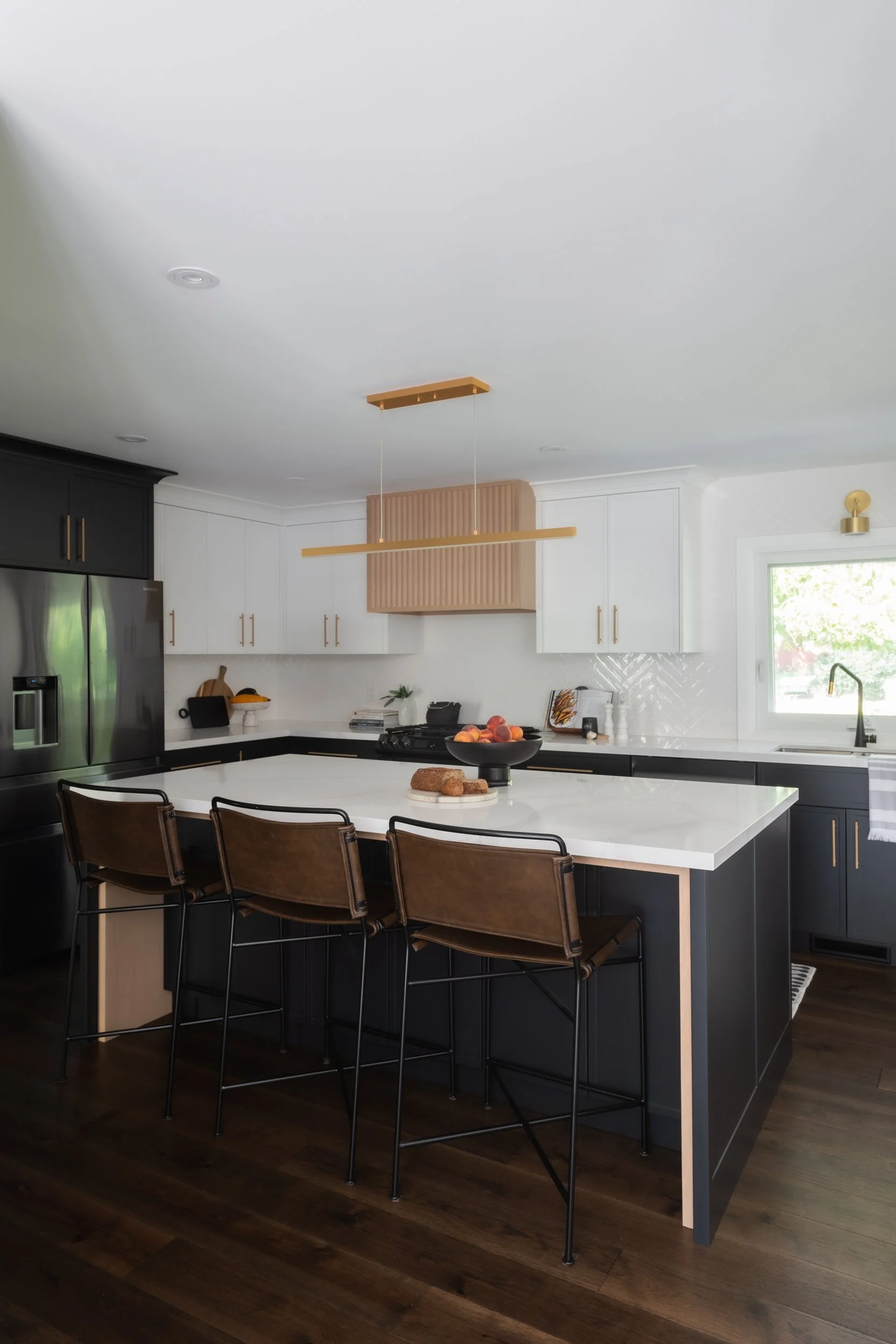Modern kitchen with black and white cabinetry, a large white kitchen island, brown leather bar stools, and stainless steel appliances near a window.