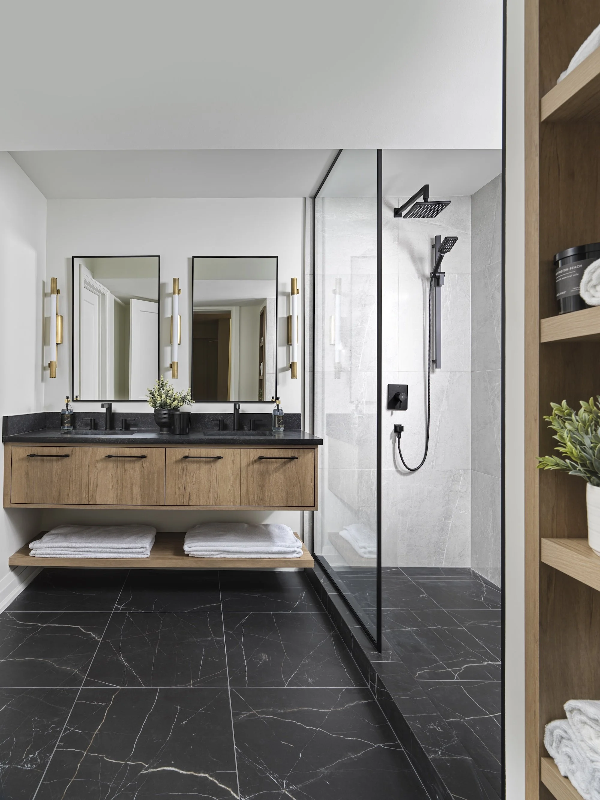 Modern bathroom with double vanity, black countertops, wood cabinets, large mirrors, and gold sconces. Walk-in shower with black fixtures and glass enclosure, black marble floor, and white walls.