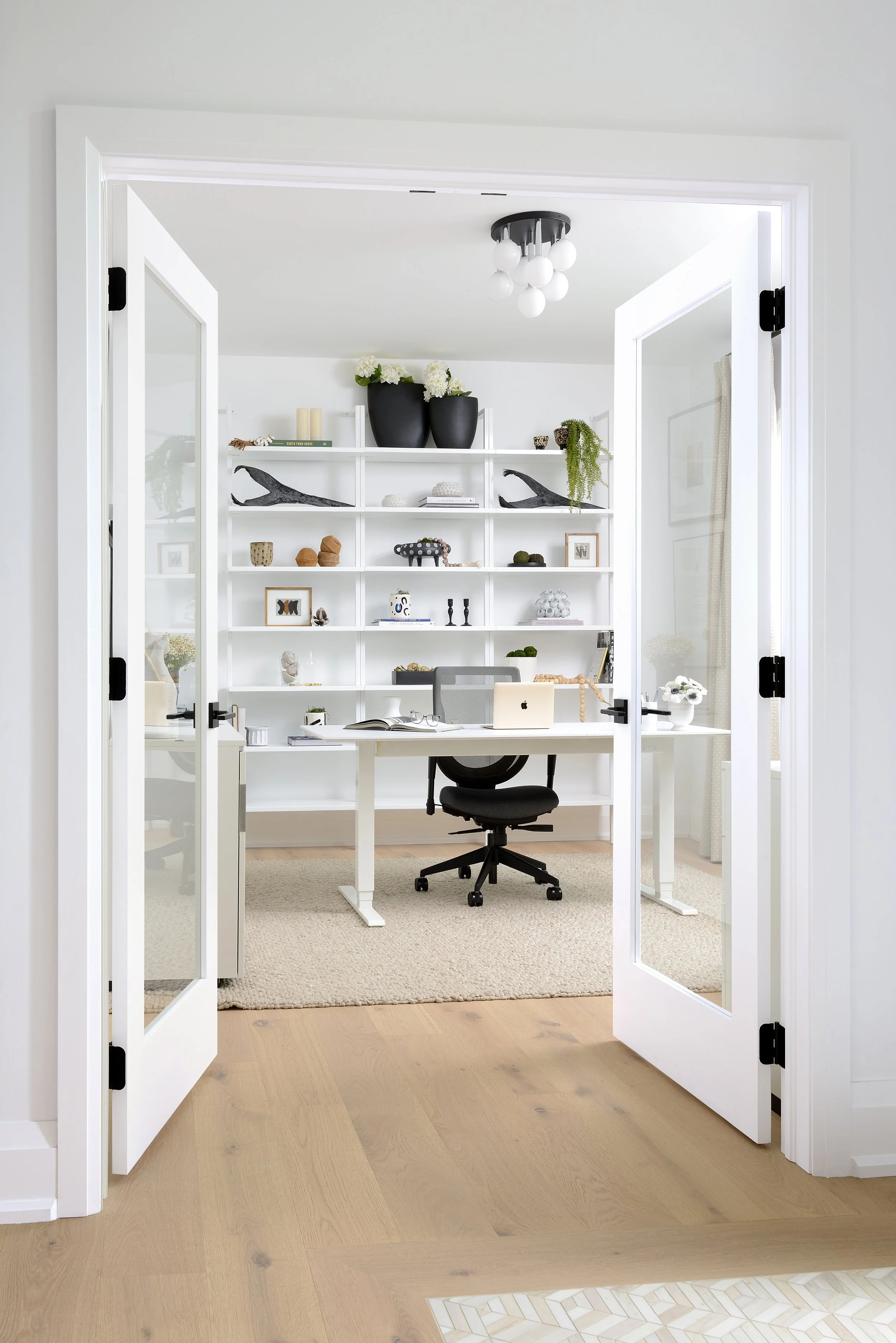 Home office with white walls, open white double doors, white desk with a MacBook, black office chair, and a white bookshelf with decorative items.