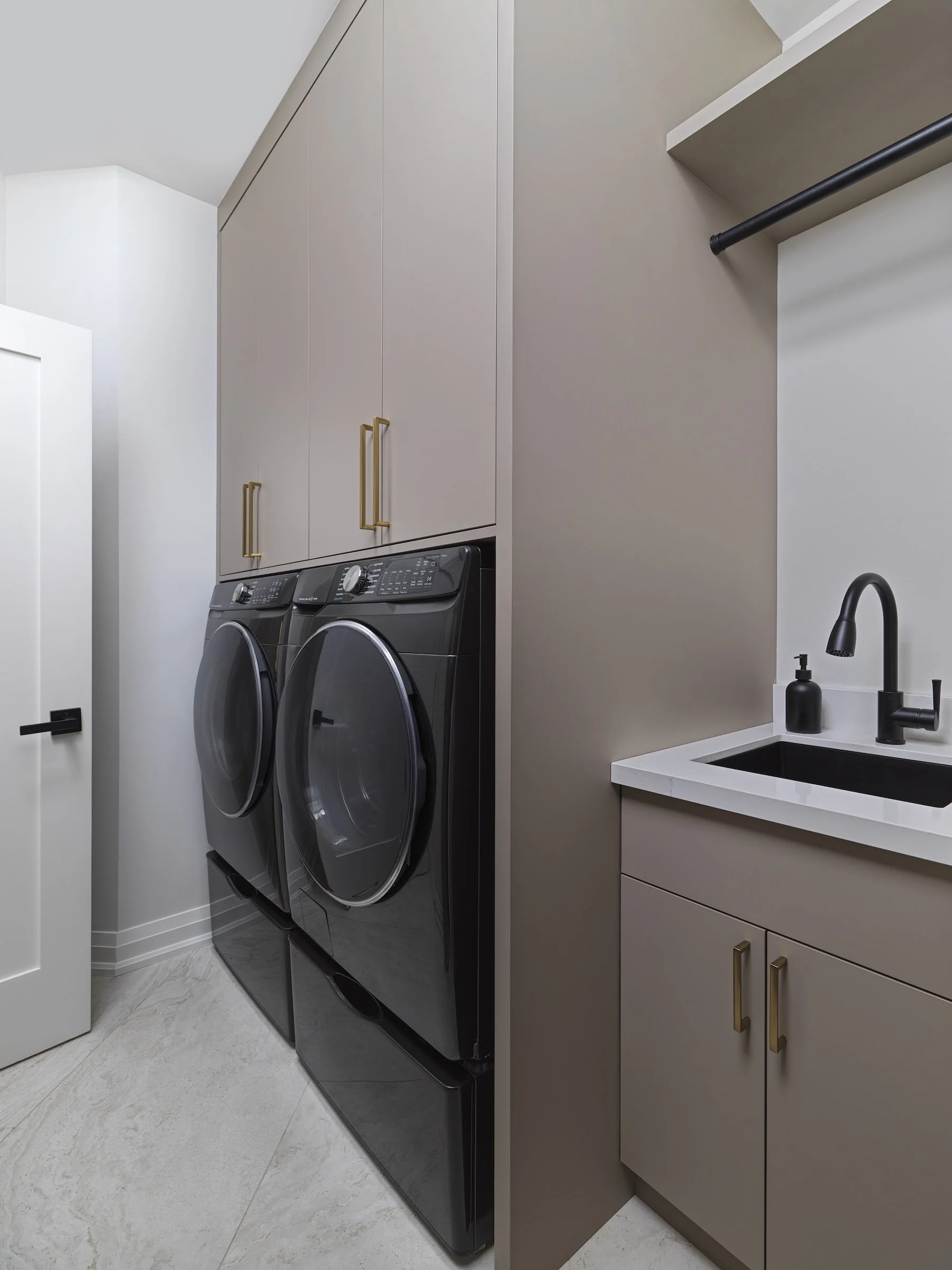 A modern laundry room with a black front-loading washer and dryer, beige cabinetry with gold handles, a white countertop with a black sink, and a black faucet.