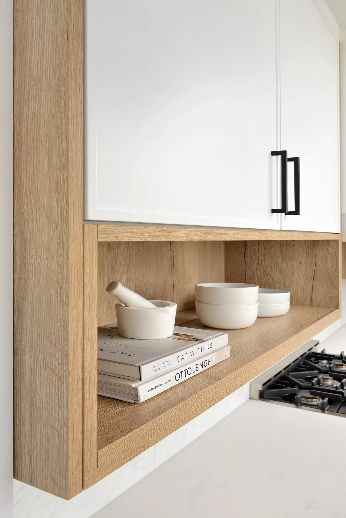 Kitchen shelf with white bowls, mortar and pestle, and cookbooks, next to a stove.