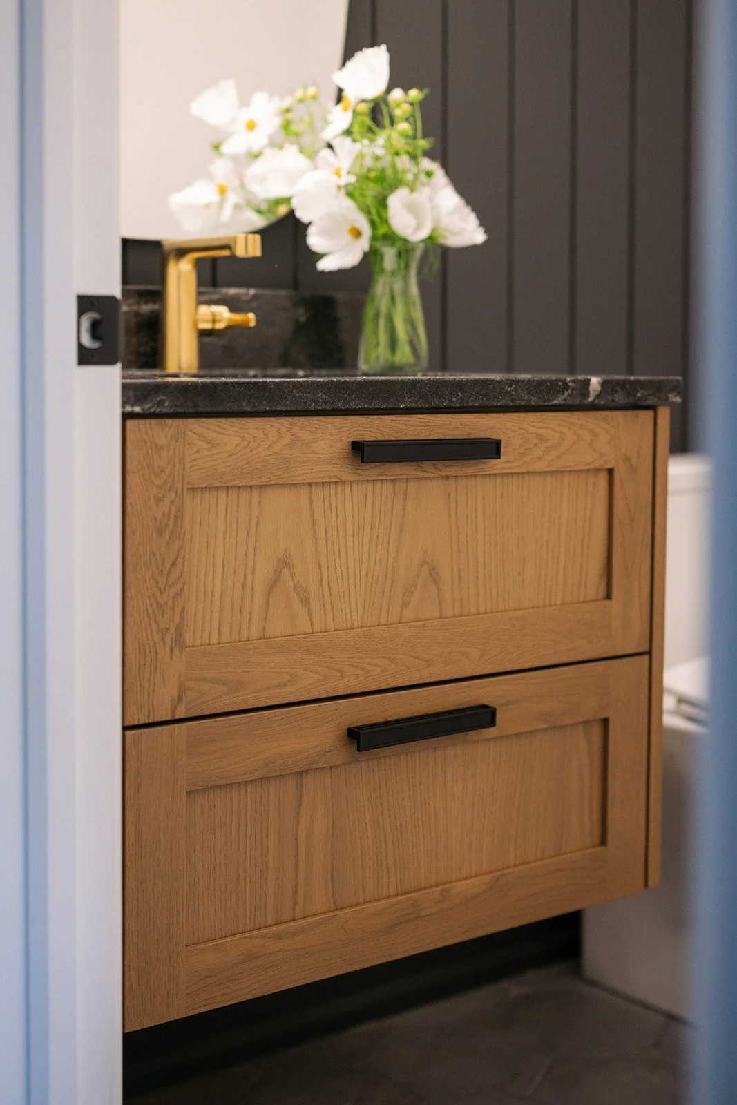 A bathroom vanity with two drawers made of light wood, featuring black handles, a black granite countertop with a gold faucet, and a vase of white flowers on the countertop. The background includes dark wood paneling on the wall.