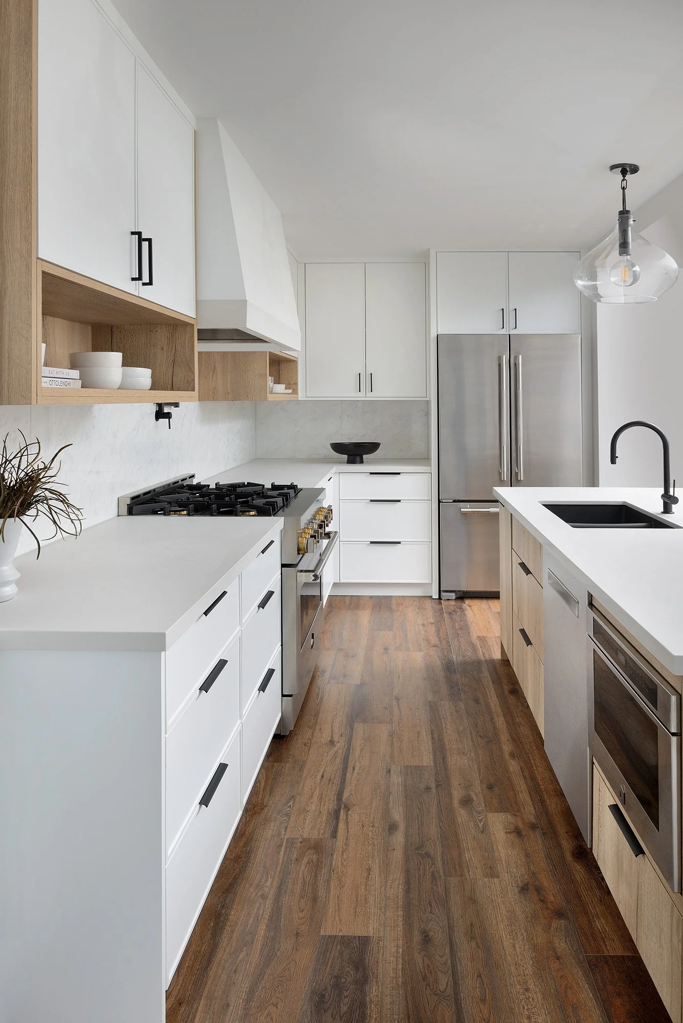 Modern kitchen with white cabinets, wooden open shelves, stainless steel refrigerator, gas stove, black sink, and hardwood flooring.